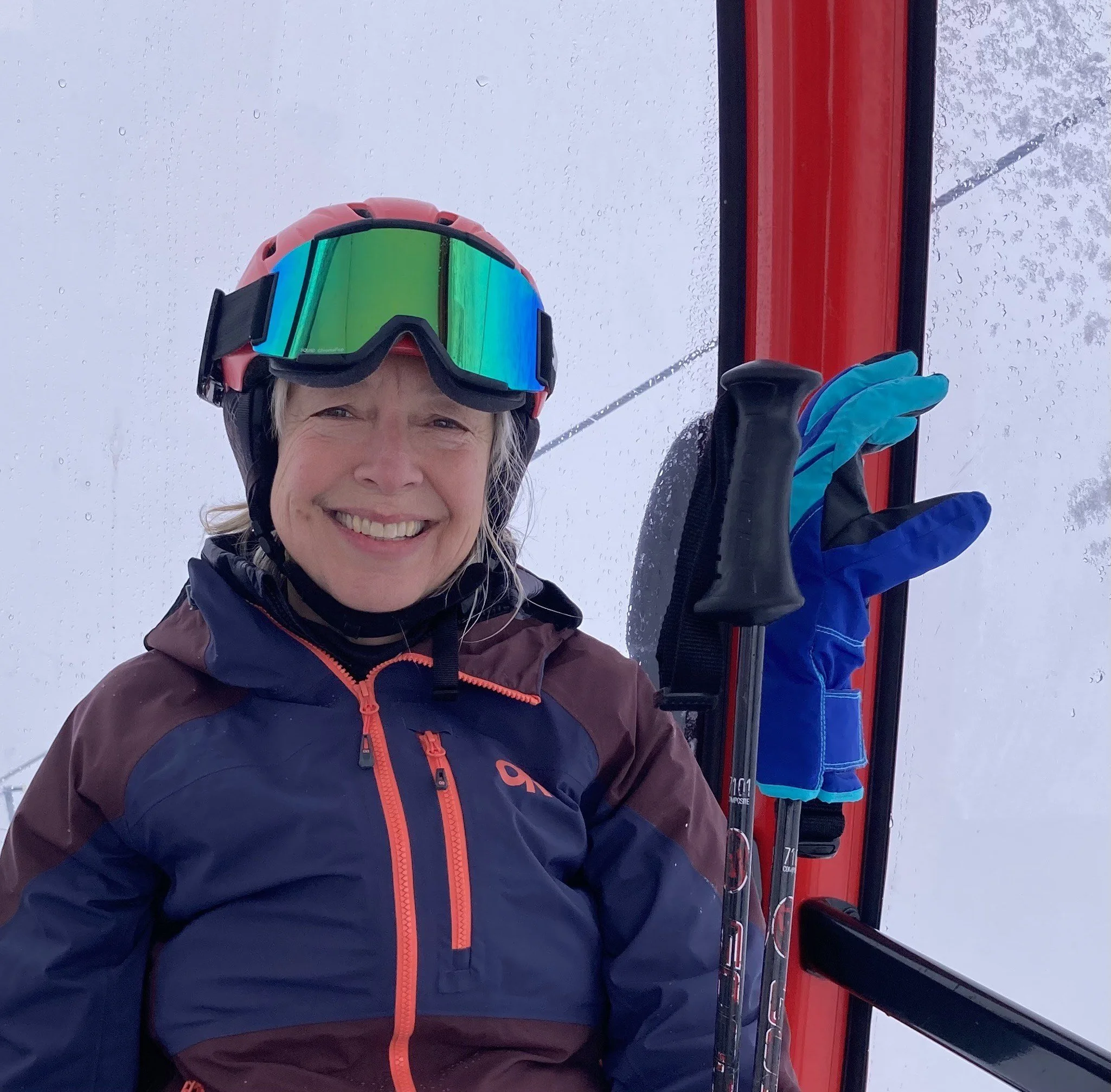 A woman in winter gear smiling on a ski lift, holding a pair of skis, with snow on the ground and snow-covered trees in the background.