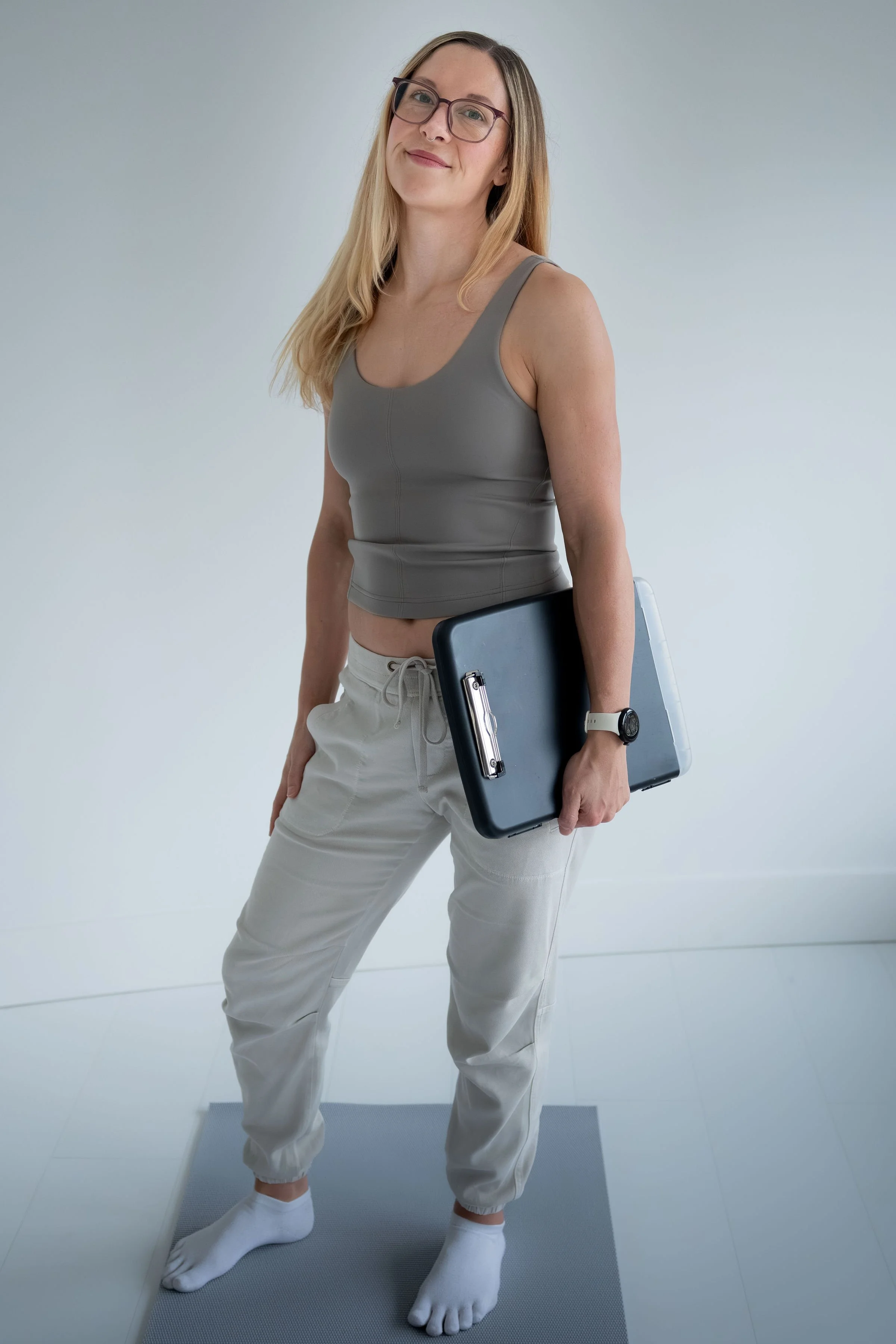 A woman with glasses and long blonde hair standing on a gray yoga mat, holding a laptop, wearing beige sweatpants, a gray tank top, and white socks, in a minimalistic room with white walls.