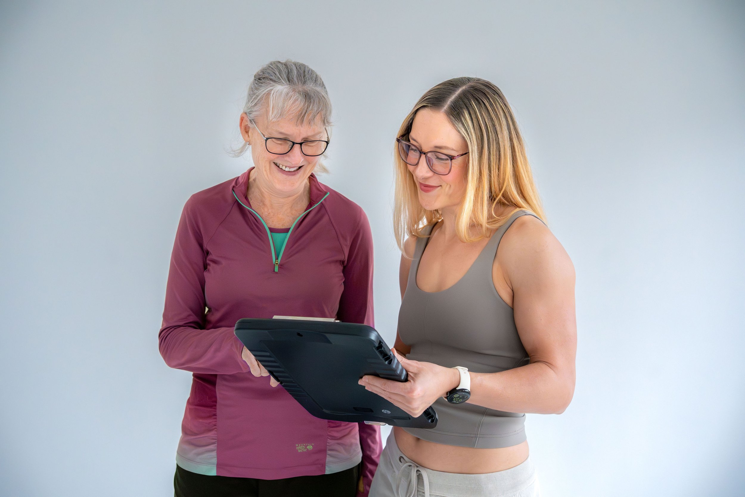 Two women, one older with glasses and gray hair, and one younger with long blonde hair and glasses, are smiling and looking at a tablet device together against a plain light gray background.