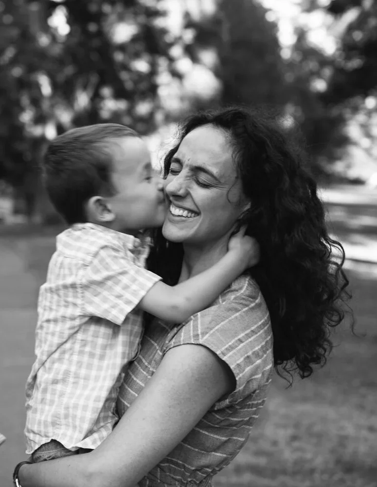 A woman with curly hair is smiling as she holds a young boy who is giving her a kiss on the cheek. They are outdoors with blurred trees in the background.