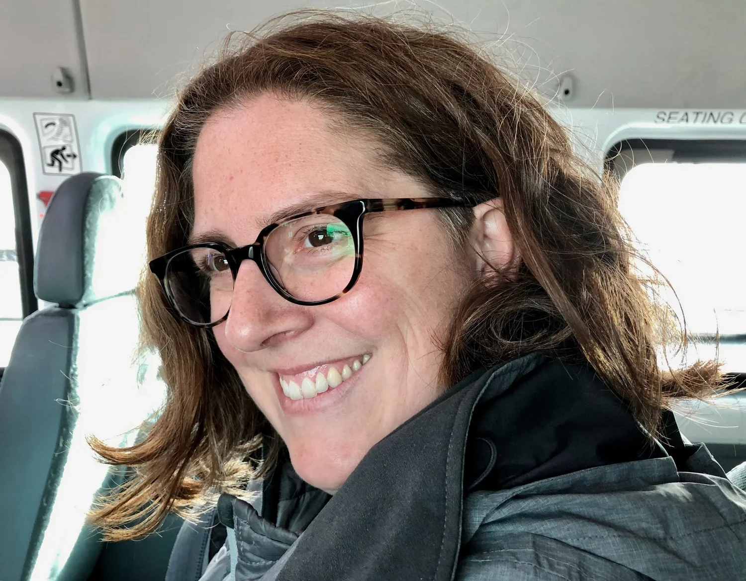 A woman with curly brown hair and glasses smiling on a bus, wearing a dark jacket.