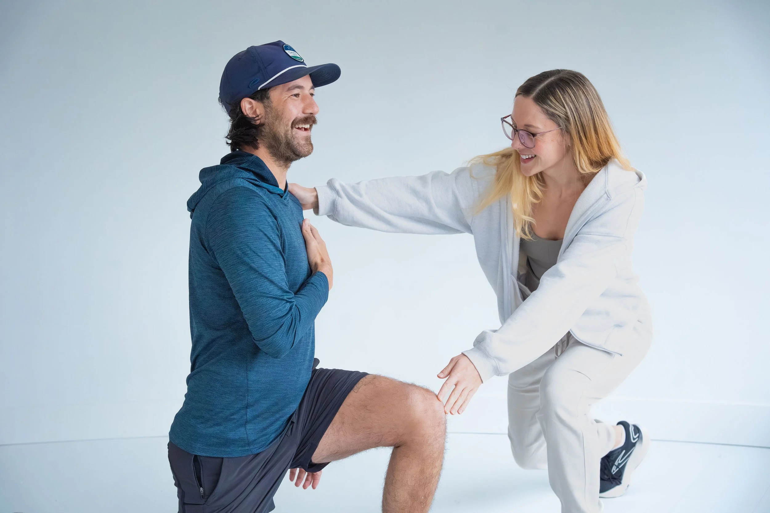 A woman with glasses and long blonde hair helping a man with a beard and a blue cap, who is kneeling, with physical therapy exercises against a plain white background.