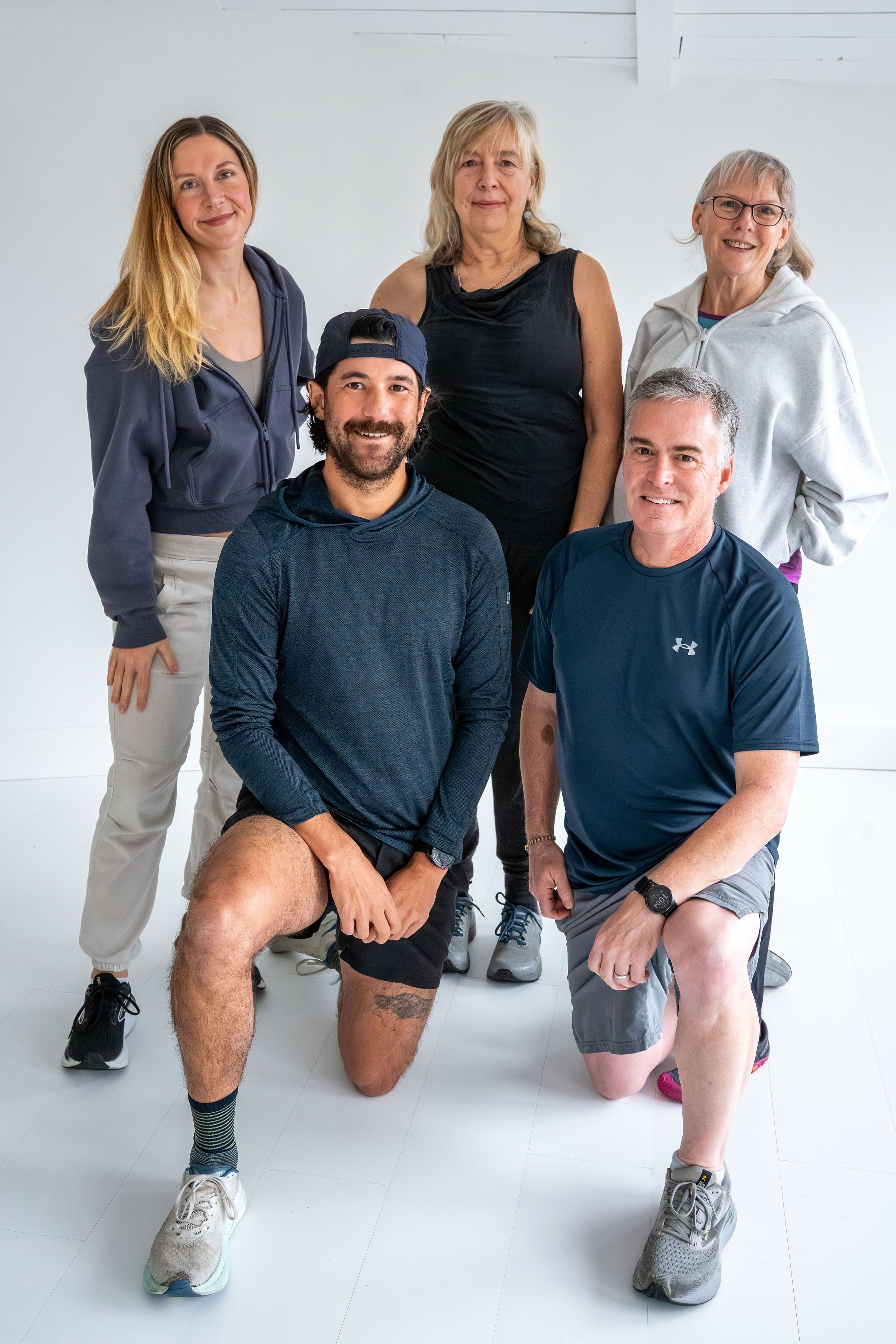 Six people, four women and two men, are posing together in a white room. Two men are kneeling in the front, one with a beard and wearing a baseball cap, and the other with short gray hair. The women are standing behind them, all smiling. They are dressed in casual athletic clothes.