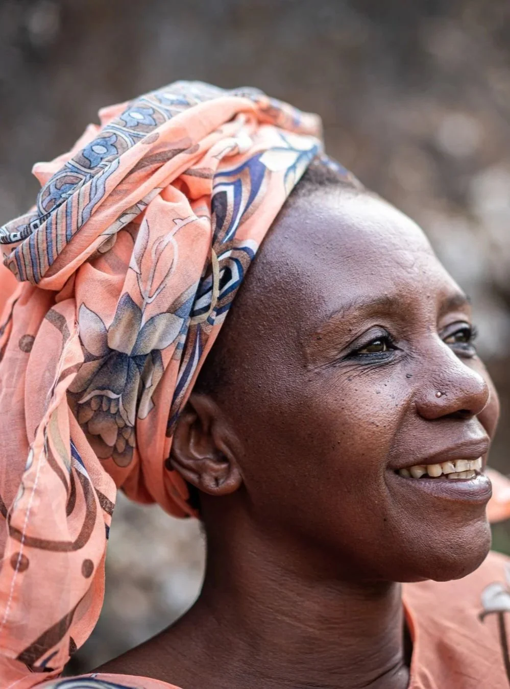 Close-up of a smiling woman wearing a colorful headscarf with floral and geometric patterns, with a nose piercing, looking to the side.