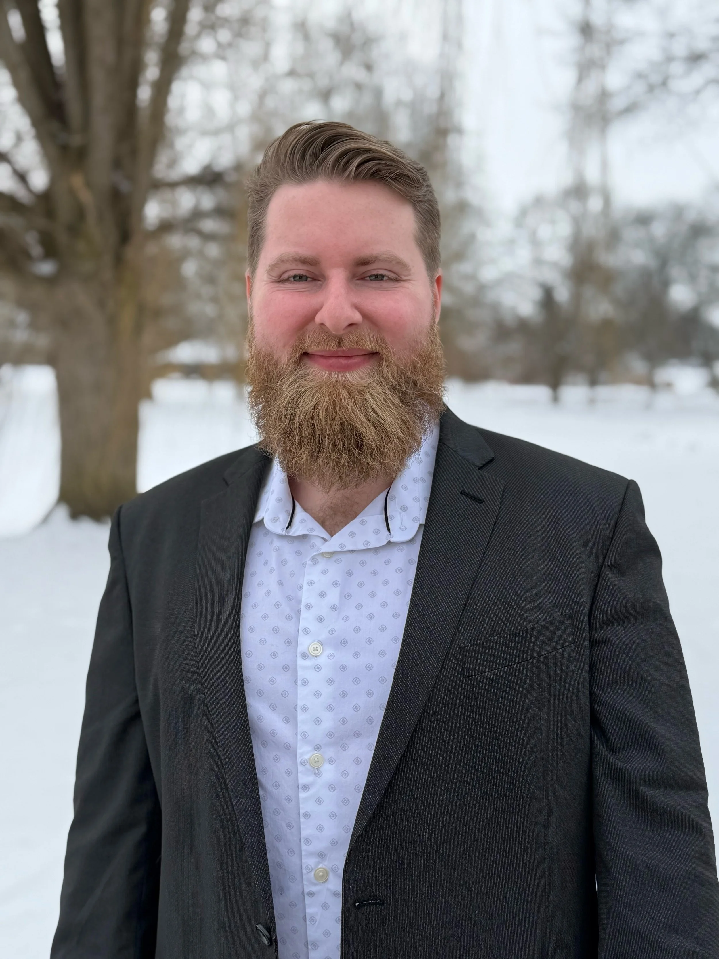 A man with a full beard and light brown hair smiling, dressed in a black suit and white patterned shirt, standing outdoors in a snowy landscape with trees in the background.