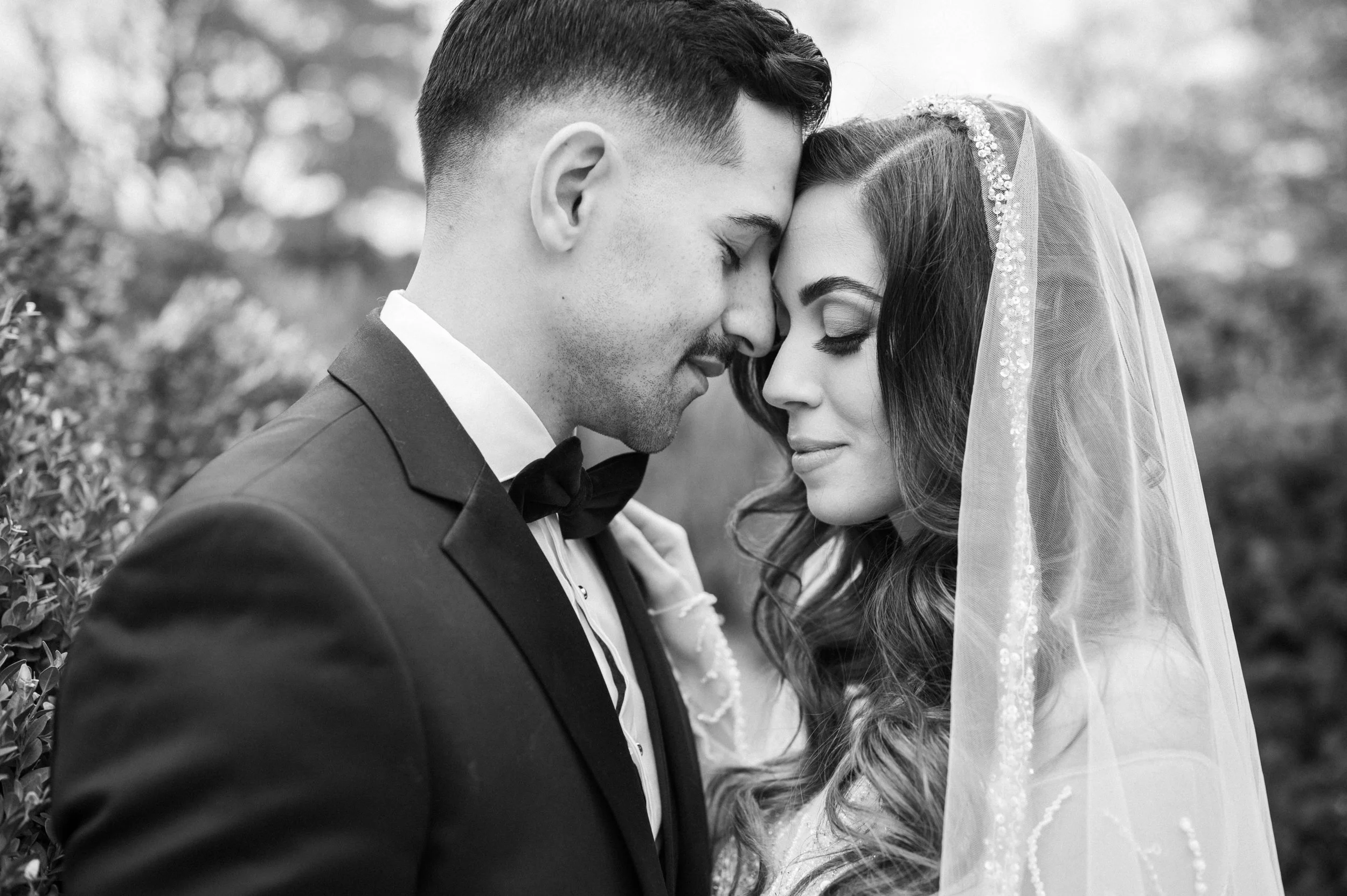 A black-and-white photo of a bride and groom close together with foreheads touching, eyes closed, outdoors.