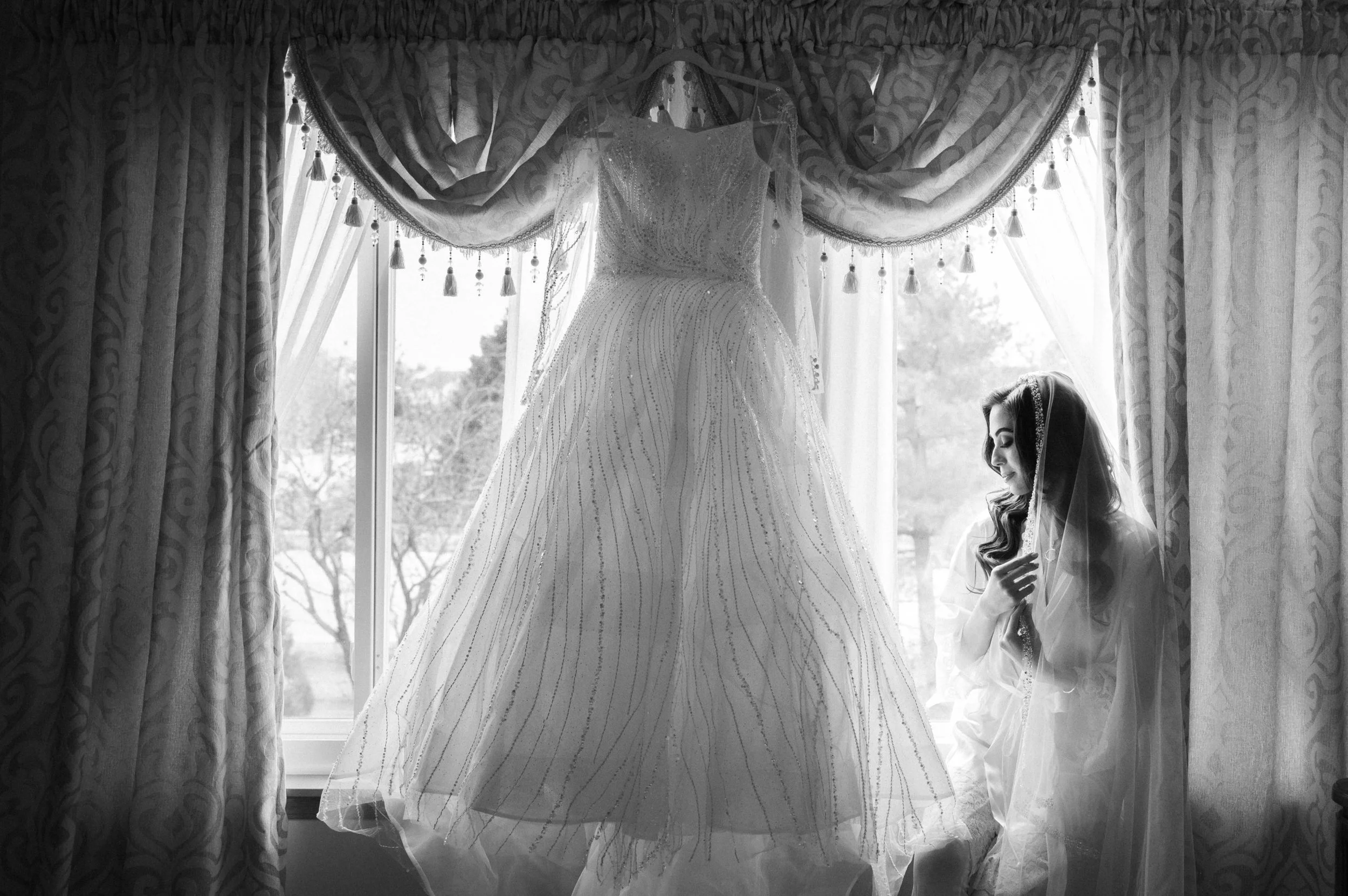 A woman in a wedding veil looks at a wedding dress hanging in a window with curtains.