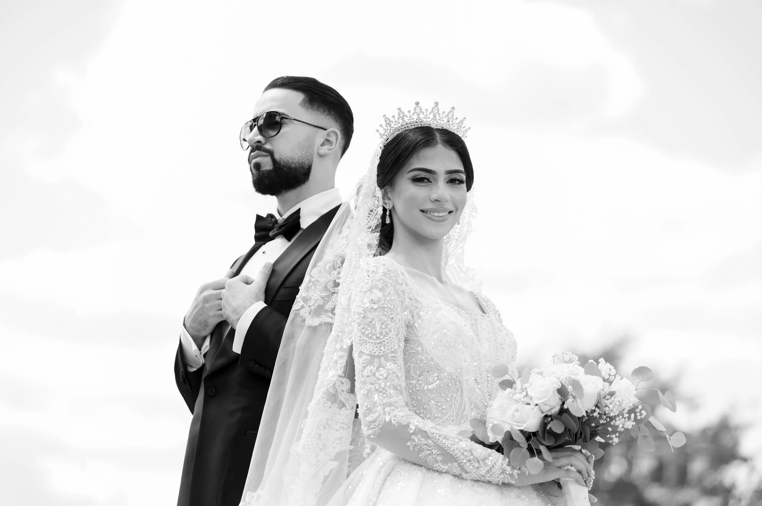 Black and white photo of a bride and groom standing back to back outdoors. The bride is smiling, wearing a lace wedding dress, veil, and a tiara, holding a bouquet. The groom is wearing a tuxedo, sunglasses, and a bow tie, with his hand on his chest.