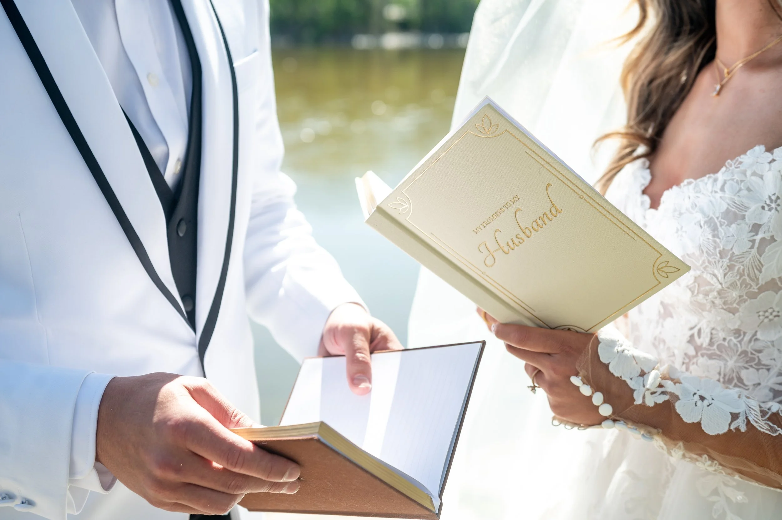 Bride and groom exchanging vows by a river, with the groom in a white tuxedo and the bride in a lace wedding dress, holding a book titled 'To My Husband'.