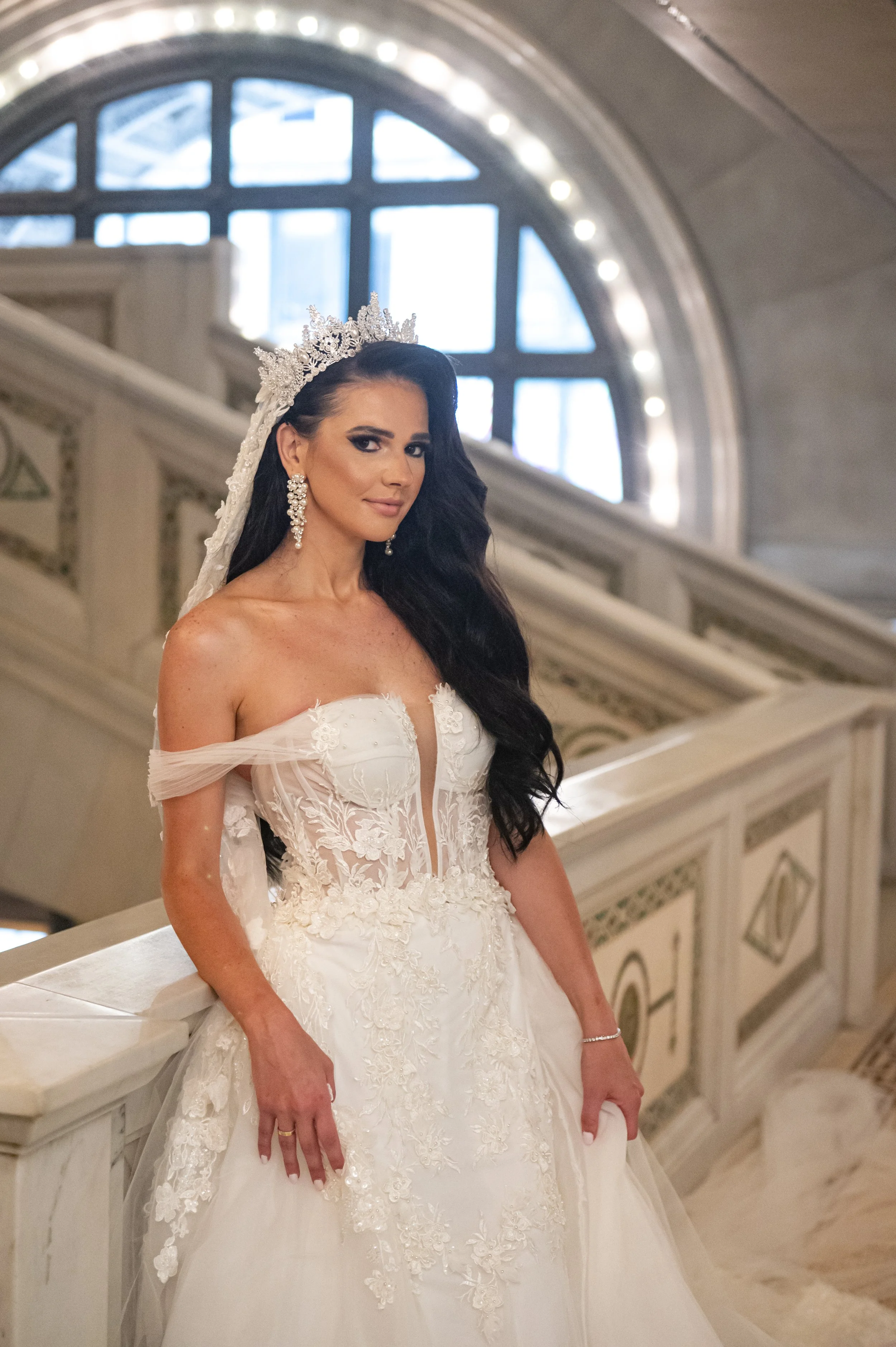 A bride in a white wedding gown with lace details, wearing a tiara and earrings, standing on a staircase inside a grand building with a large arched window in the background.