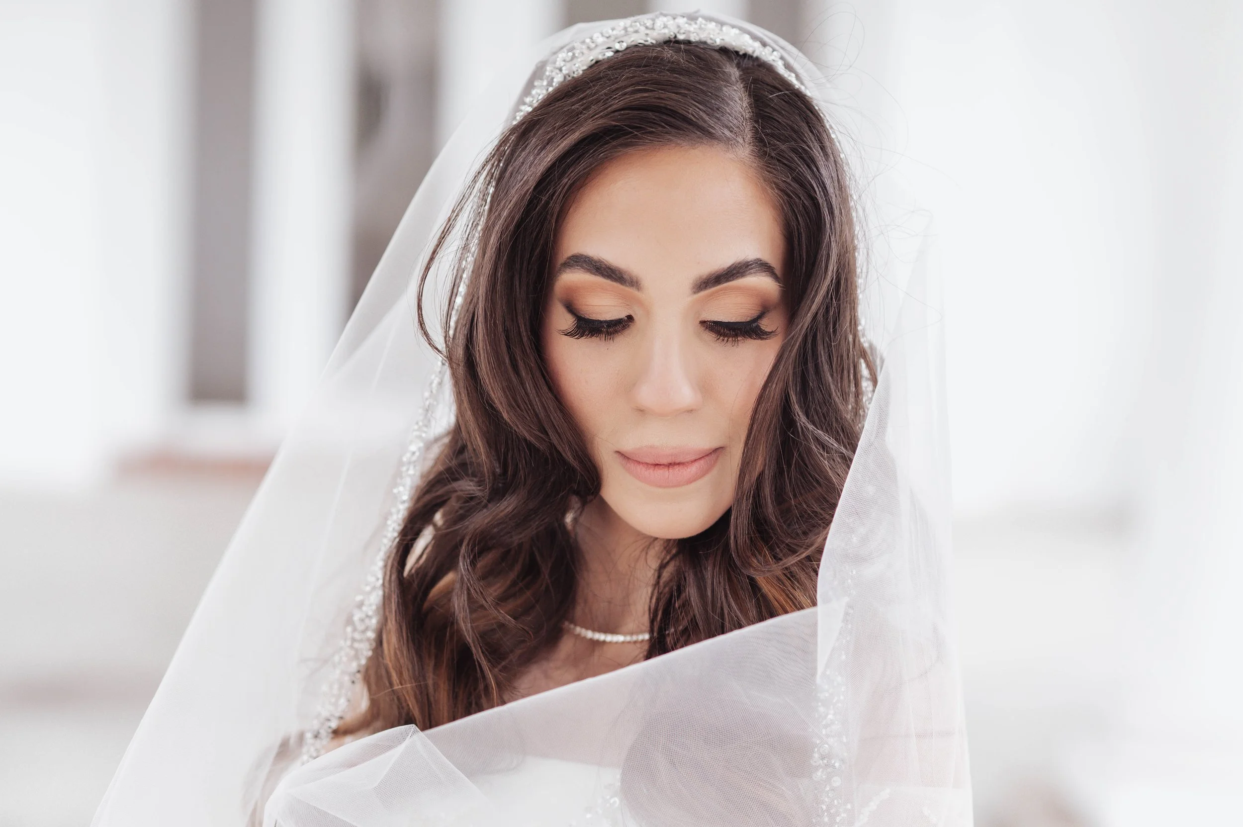 Close-up of a bride with long wavy brown hair, wearing a pearl necklace, with a veil over her head, and her eyes closed.