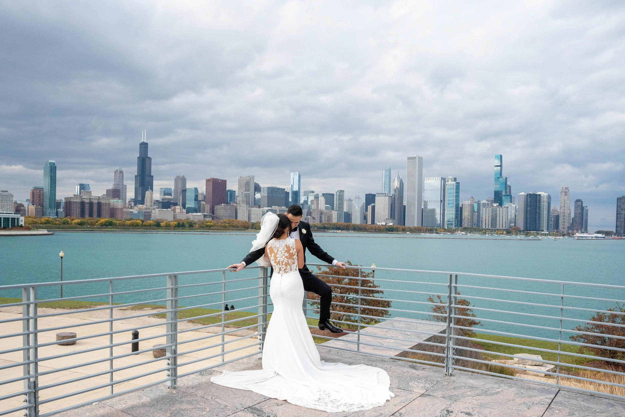 A bride and groom standing on a viewing platform with a city skyline and lake in the background, during a cloudy day.