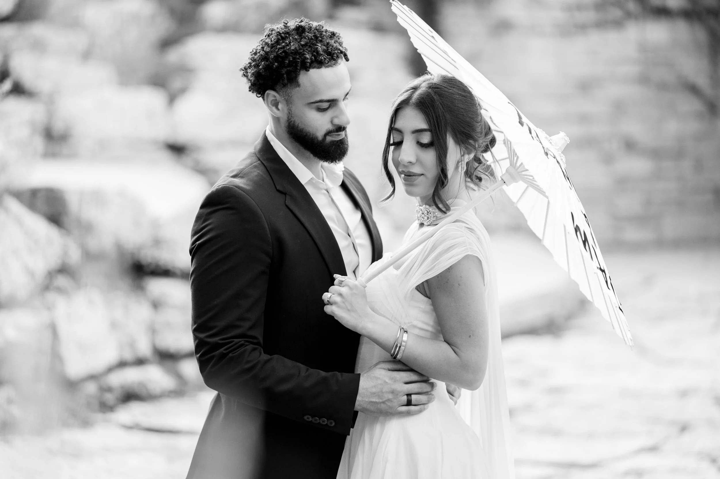 A black and white photo of a couple embracing outdoors. The man has curly hair and a beard, wearing a suit. The woman has long hair and is holding an umbrella, wearing a dress.