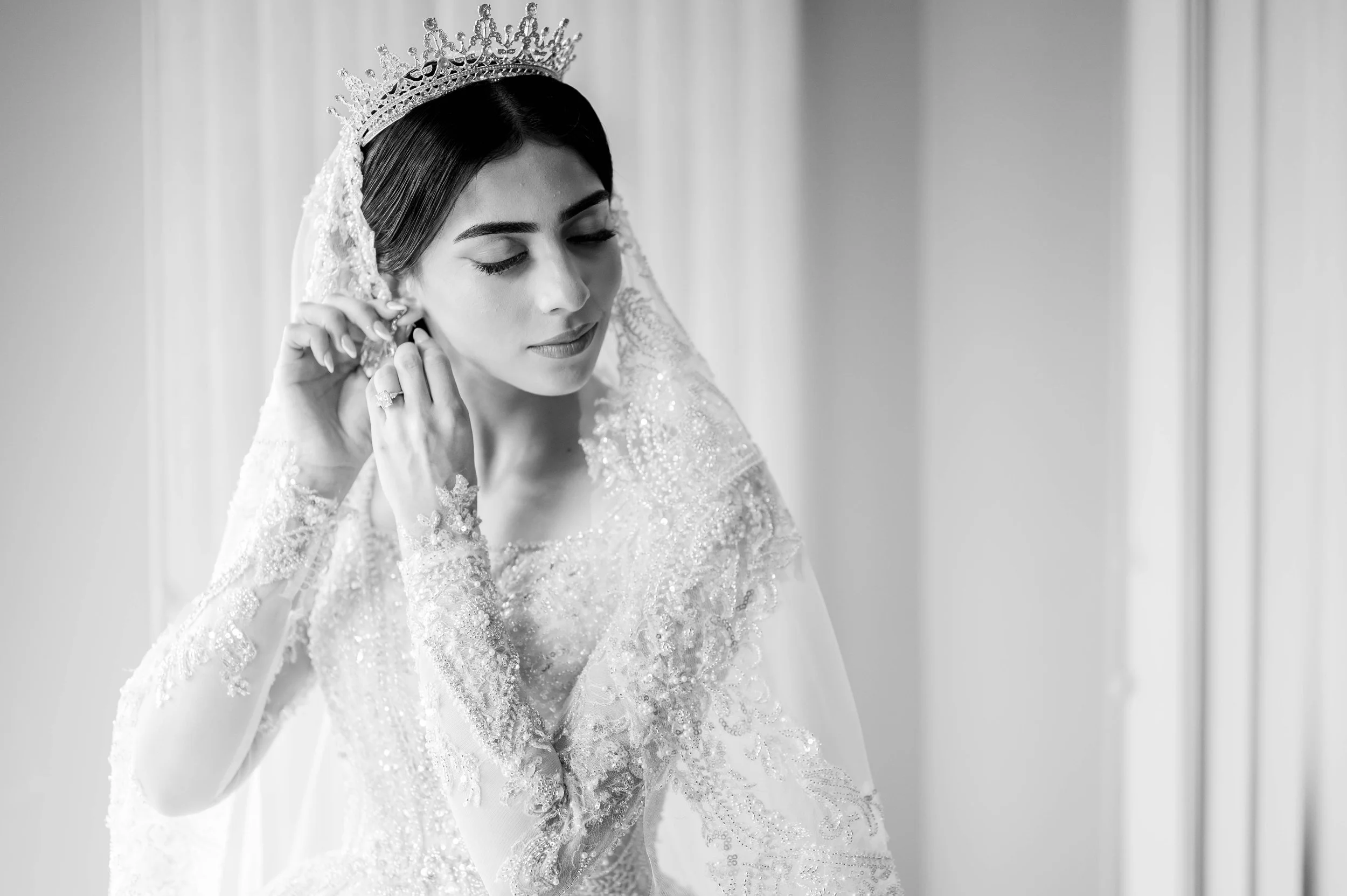 Black and white photo of a bride adjusting her earring, wearing a bejeweled gown and a tiara, with a serene expression and eyes closed.