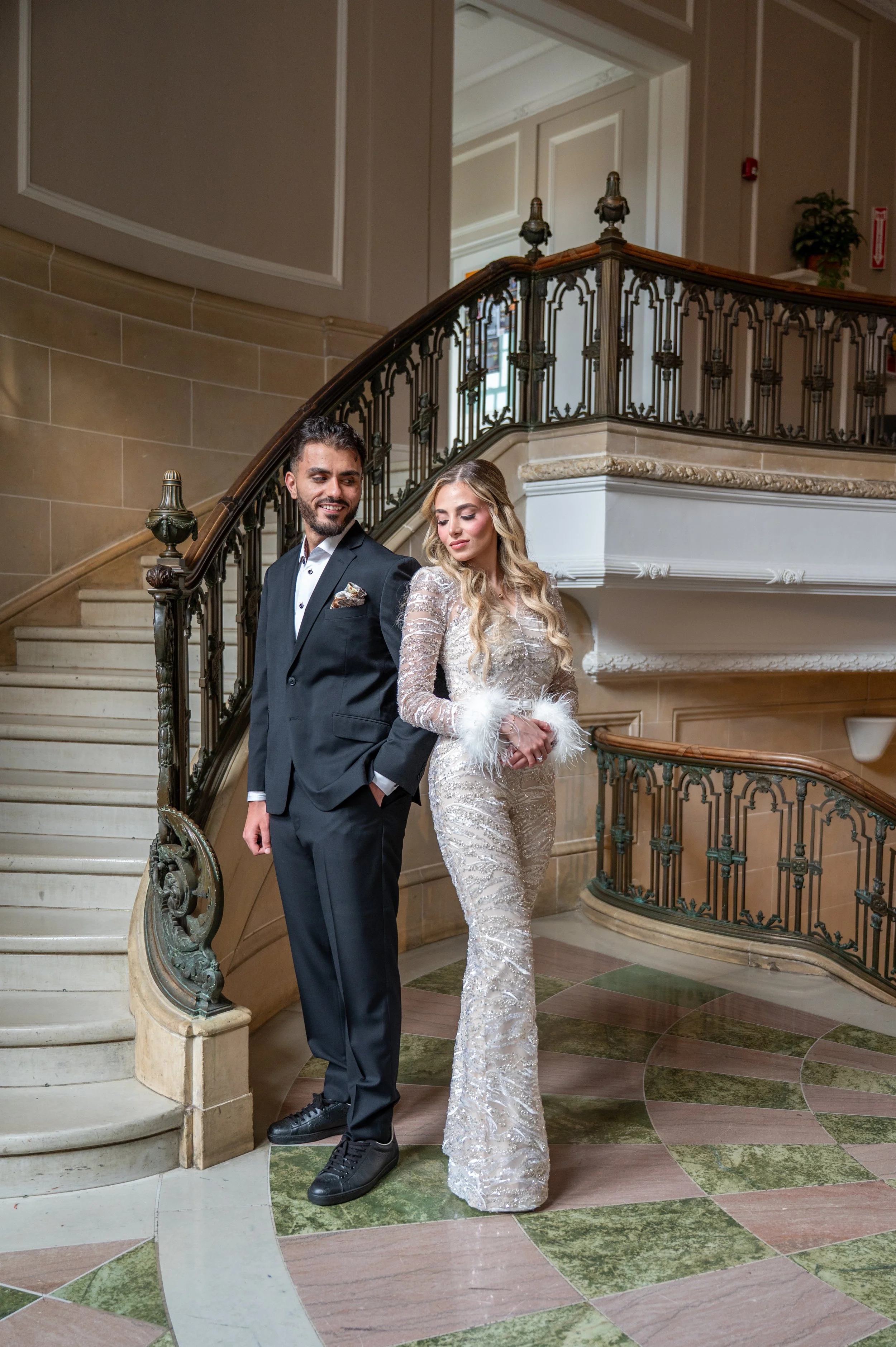 A well-dressed couple standing on a staircase in an elegant interior, the man in a black suit with a white shirt and the woman in a glamorous silver sequined gown with feathered cuffs.