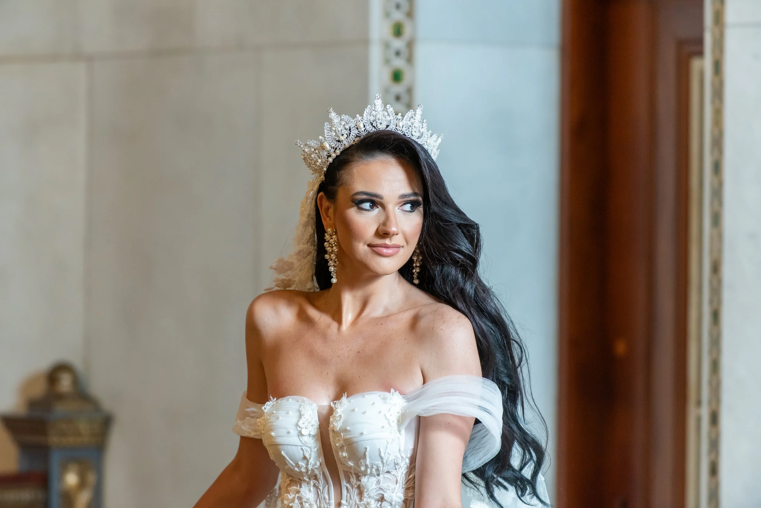 A woman with long black hair wearing a white off-shoulder wedding dress, elaborate earrings, and a large crown, standing indoors near a window.