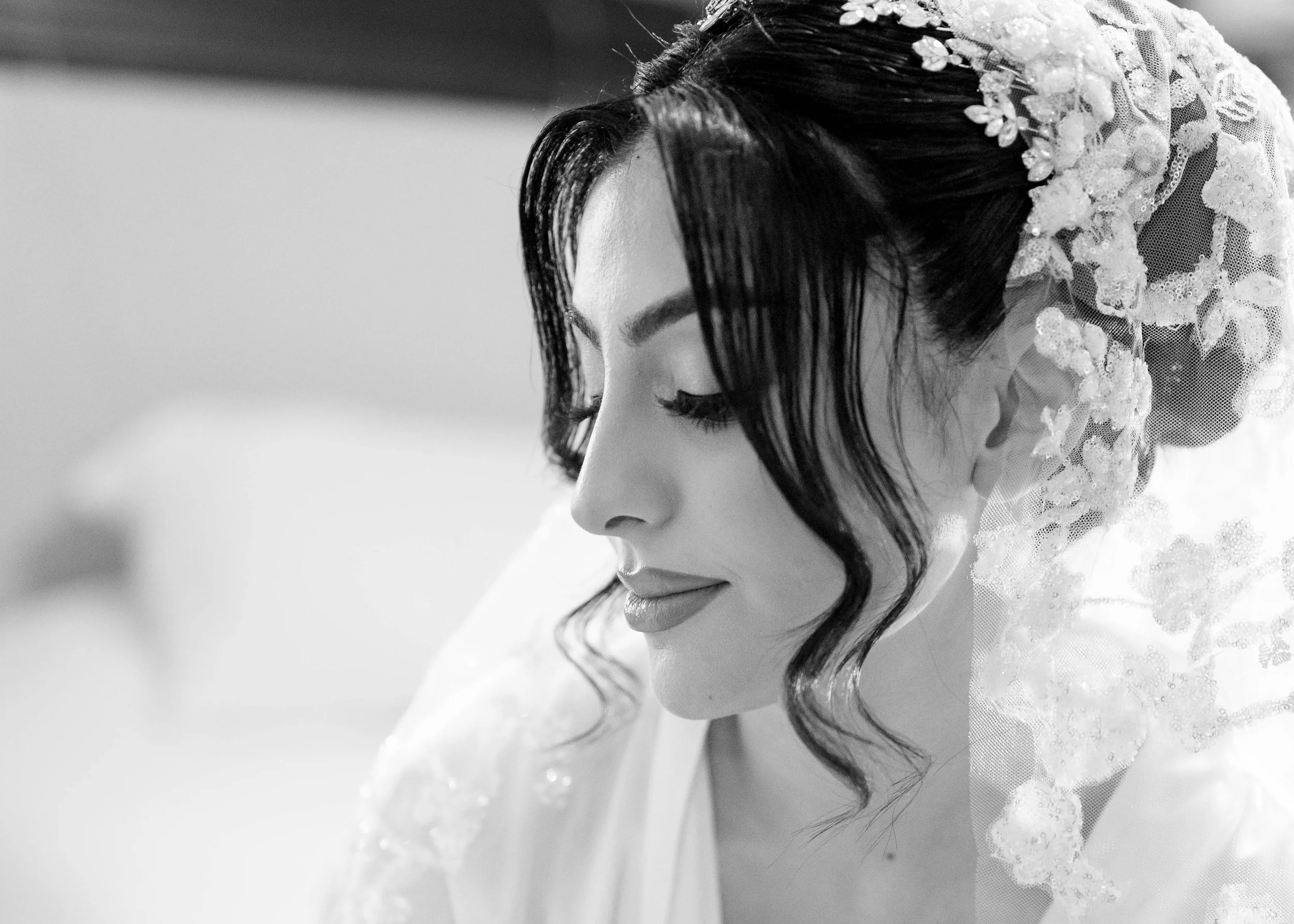 Black and white photo of a woman with dark hair styled in loose waves, wearing a delicate lace veil and a white garment, with a serene expression and eyes closed.