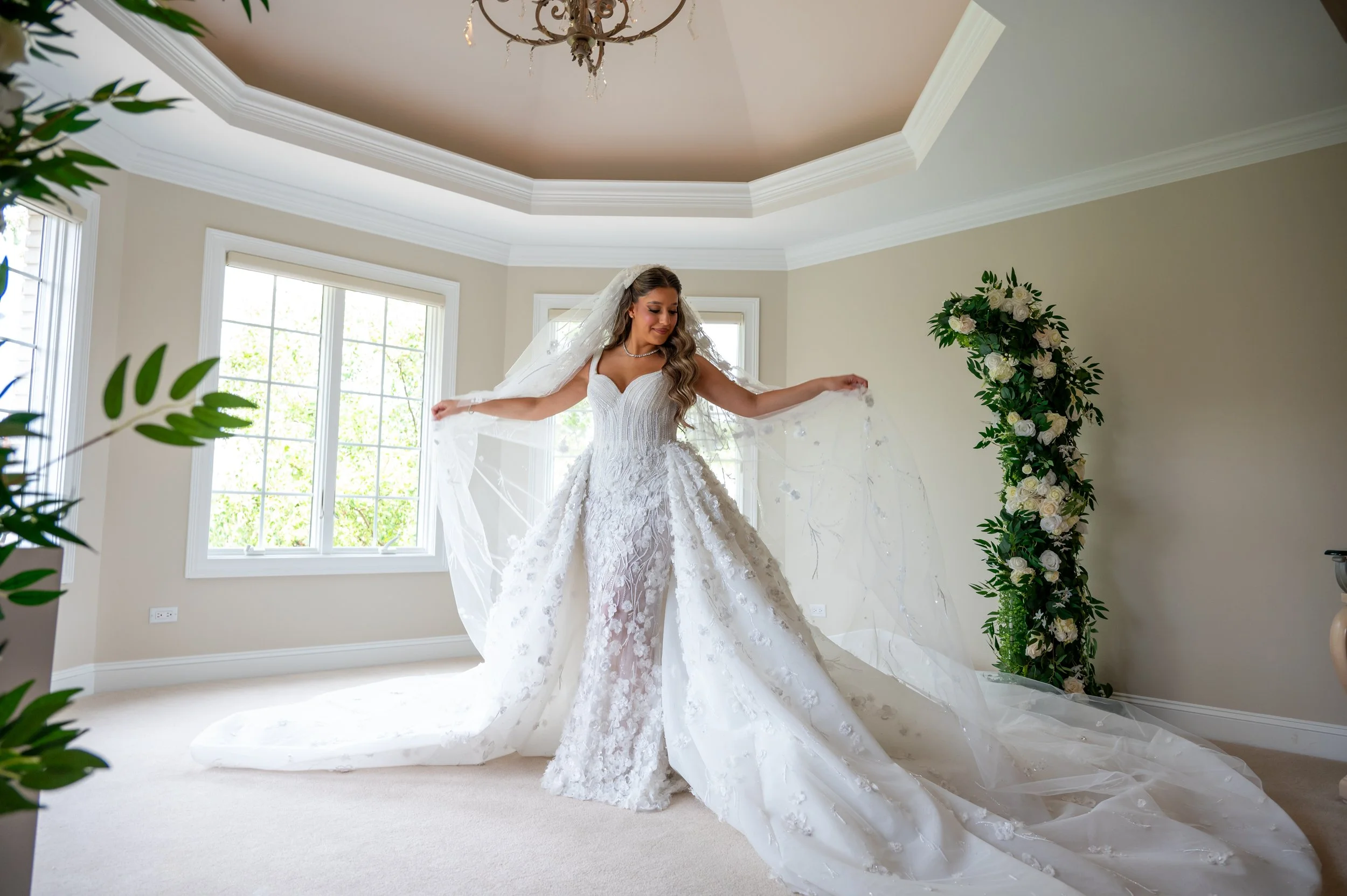 Bride in a wedding dress with a long train, standing in a bright room with large windows, floral arch on the side, and chandelier above.