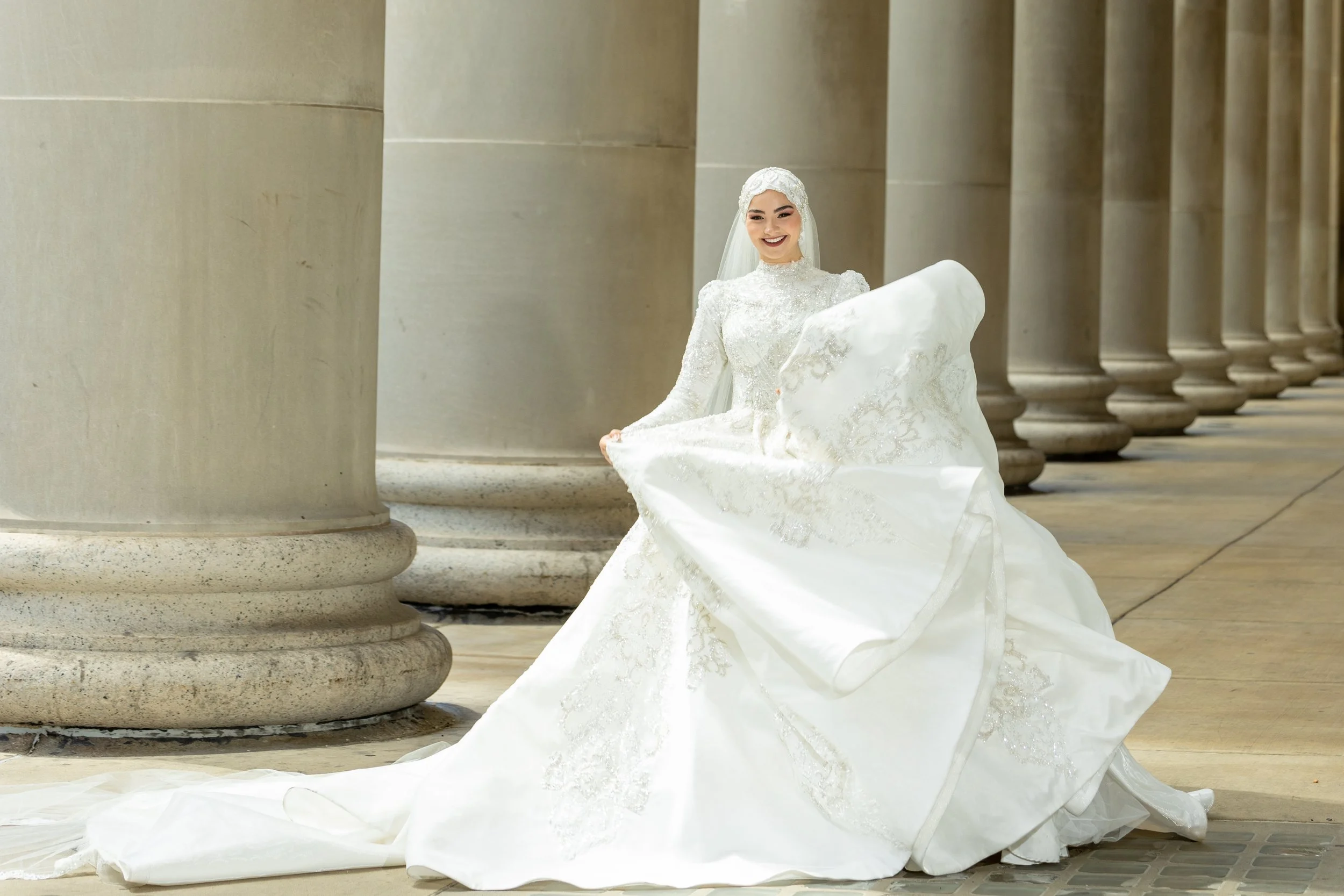 A bride in a white wedding dress with intricate embroidery, holding the skirt of her gown, standing among large stone columns.