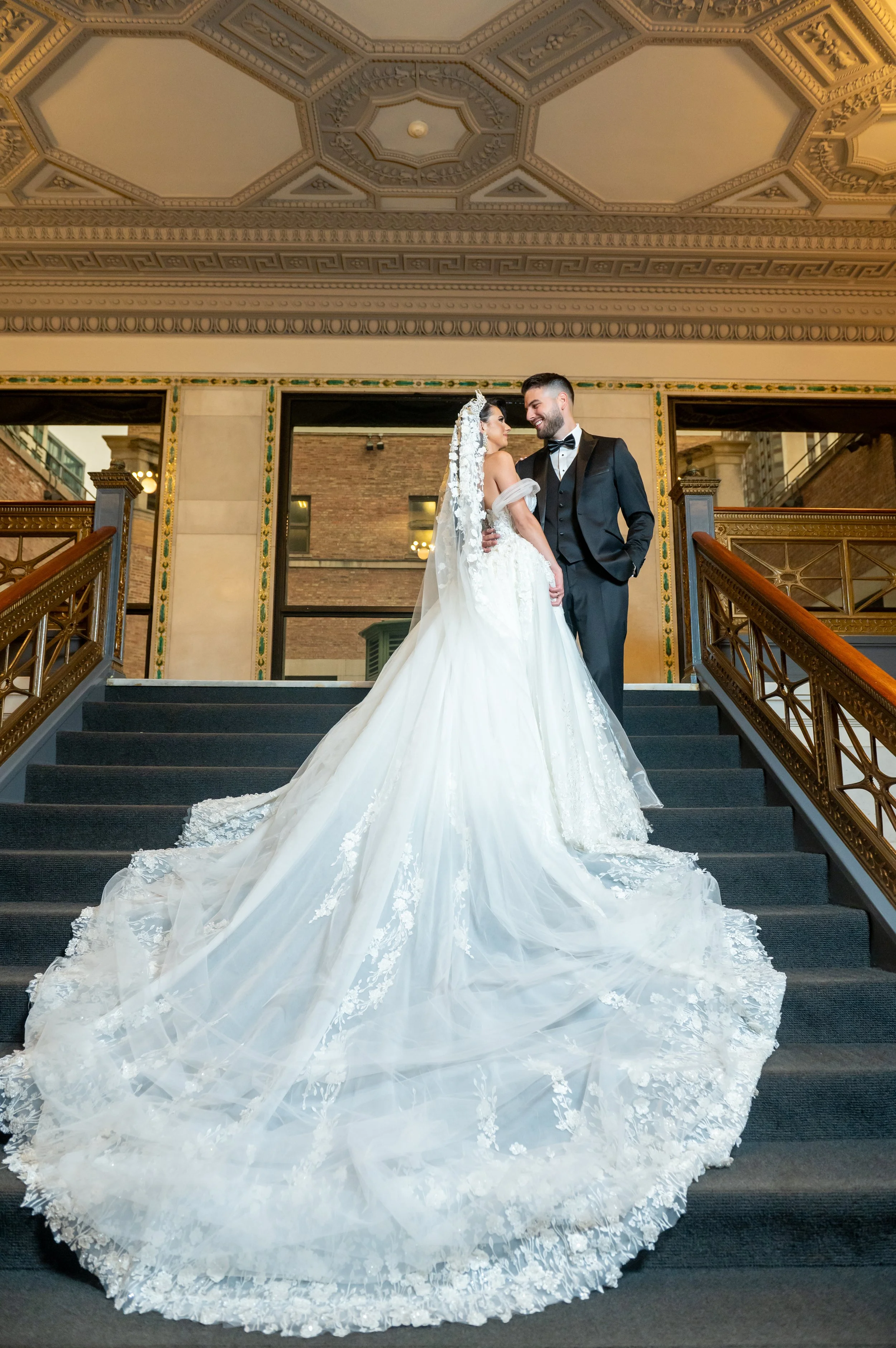 A bride and groom standing on a staircase, smiling at each other, with the bride wearing a long, flowing white wedding gown and veil, and the groom in a black tuxedo, inside a historic building with ornate ceiling details.