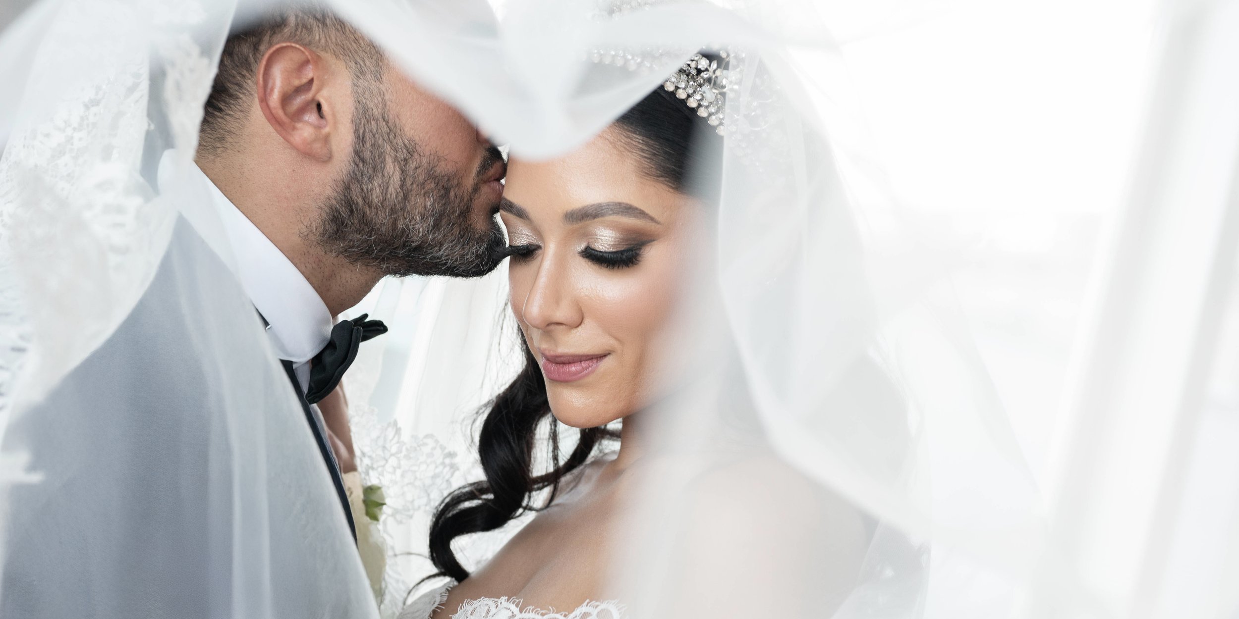 A bride and groom sharing an intimate moment under the bride's veil, with the groom kissing the bride's forehead, both with their eyes closed.