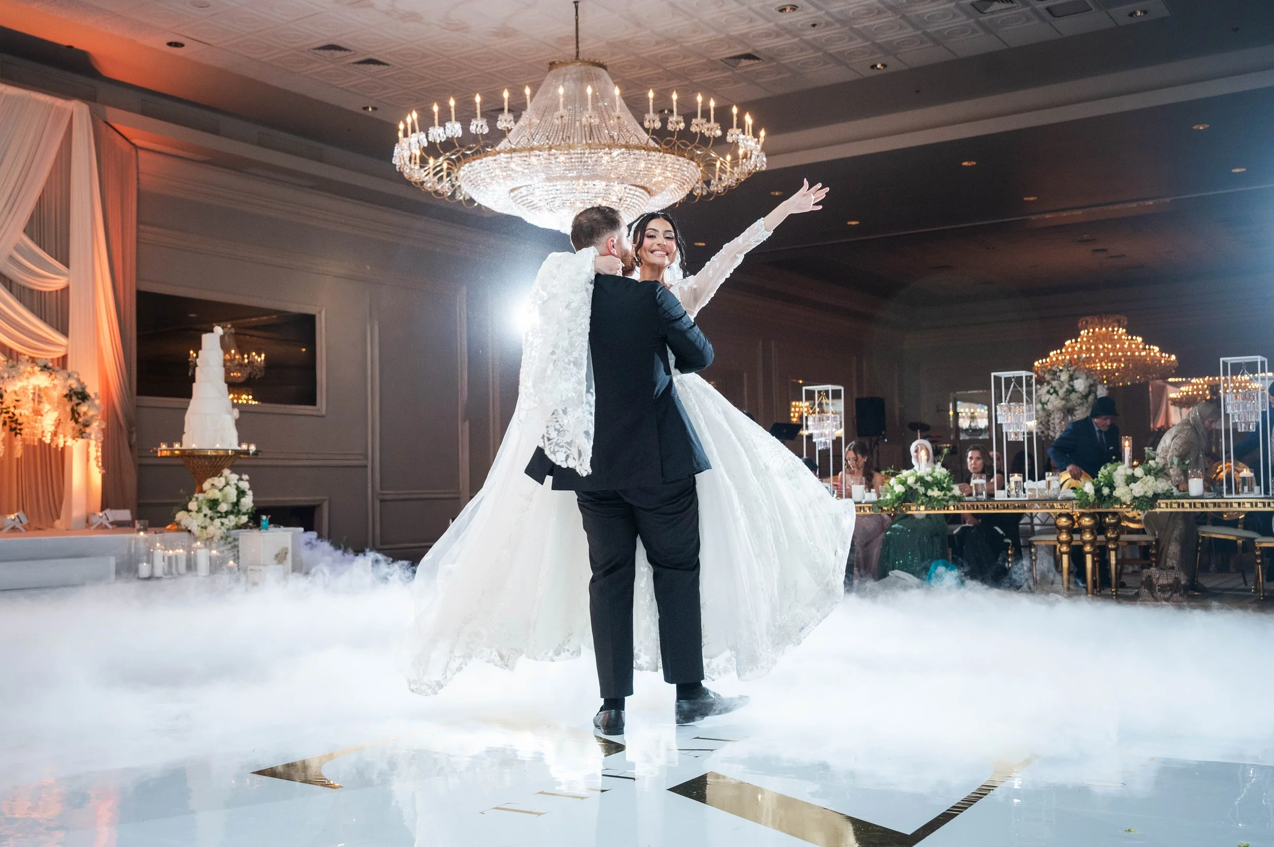 A bride and groom dancing on a decorated wedding reception dance floor, with a chandelier overhead and guests seated at tables in the background.