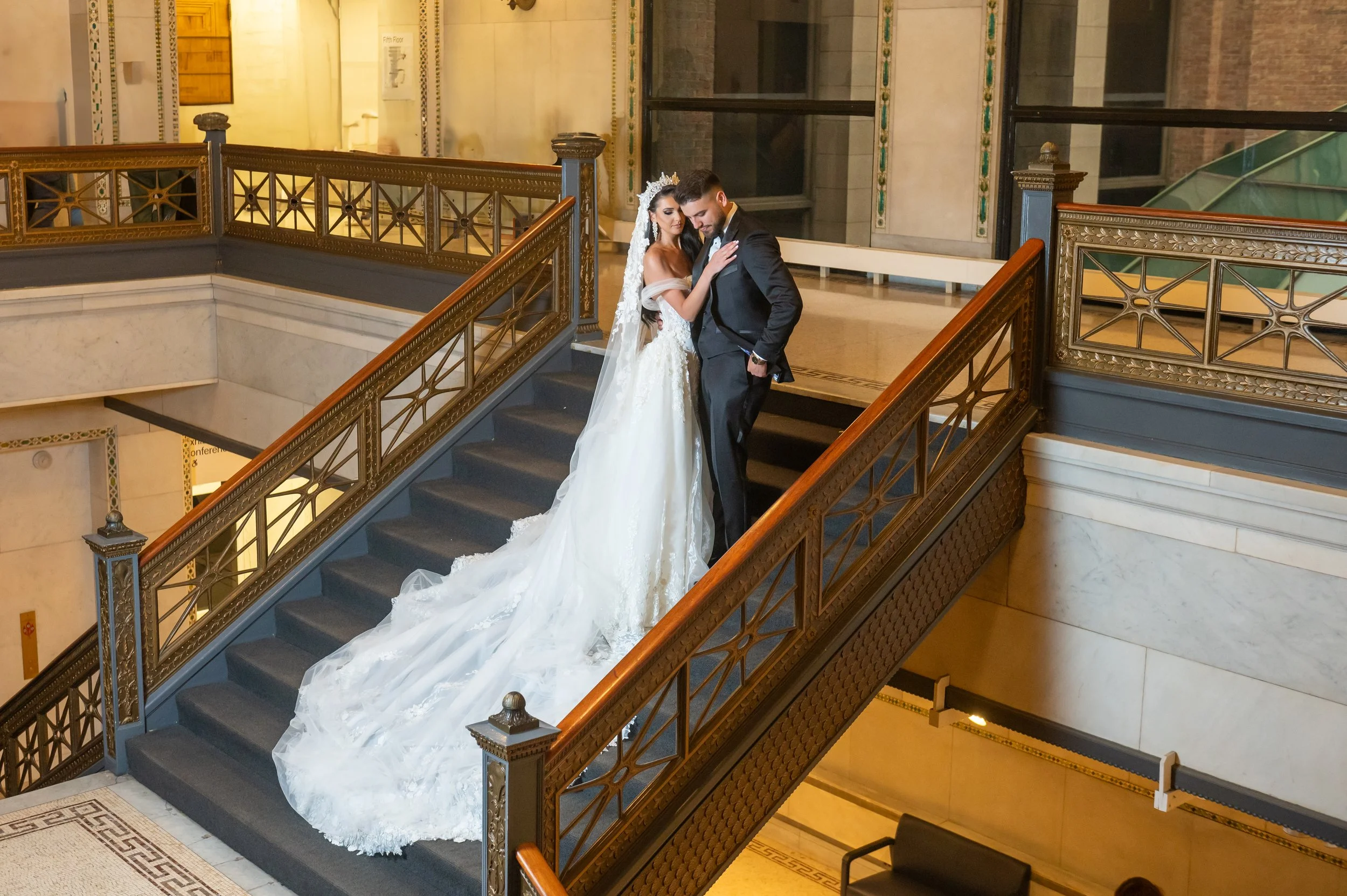 A bride and groom in wedding attire standing on a staircase inside a grand building, embracing each other.