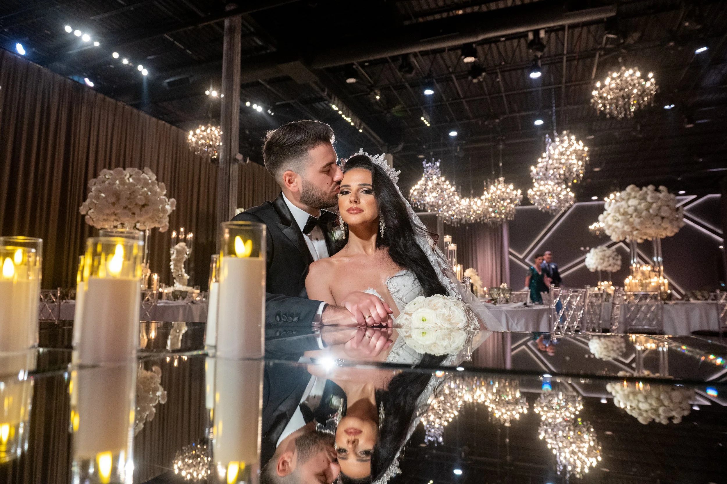 A bride and groom sitting at a wedding reception table with elegant chandeliers and floral centerpieces in the background.