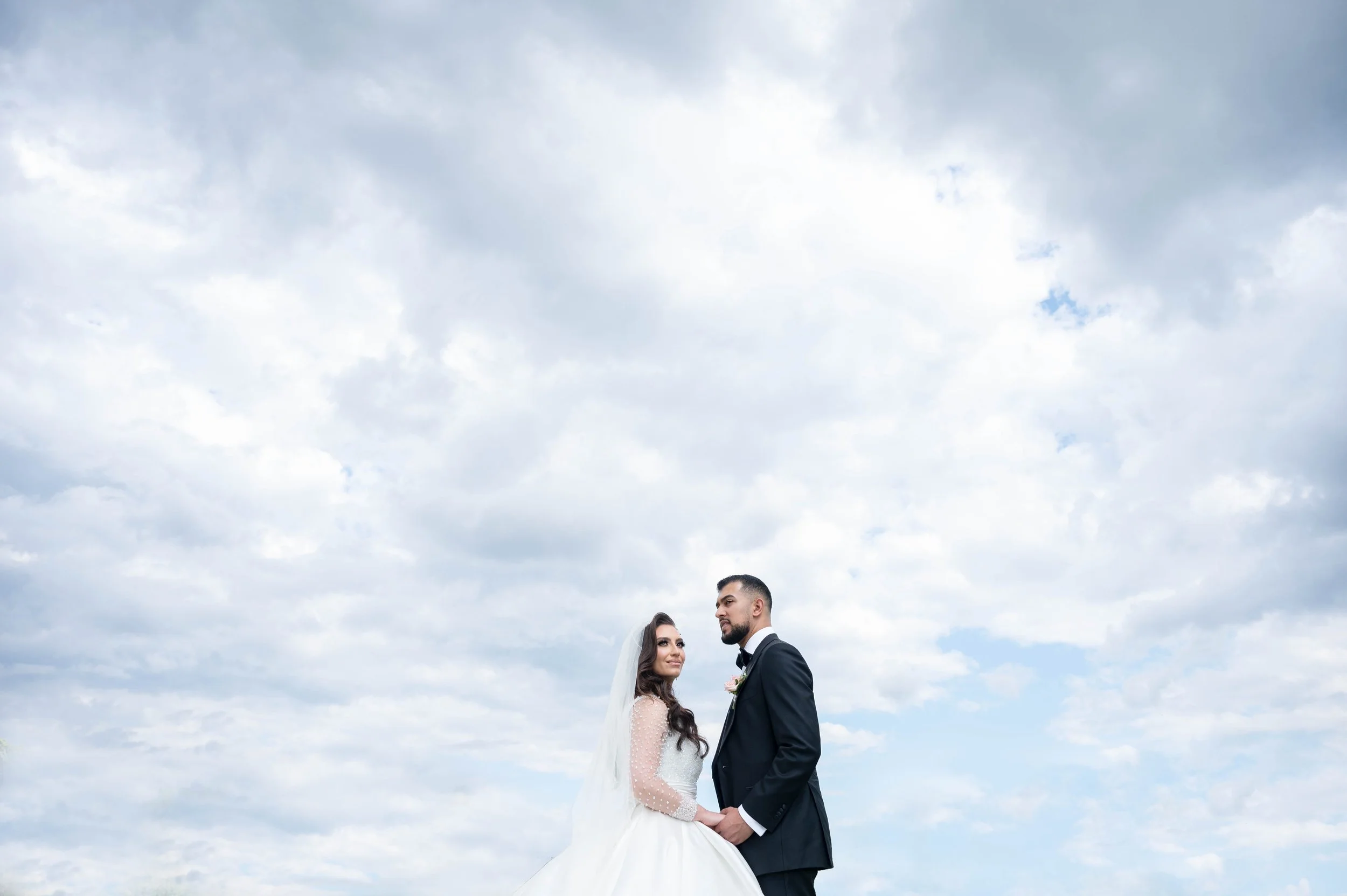 Bride and groom holding hands outdoors under cloudy sky on their wedding day.