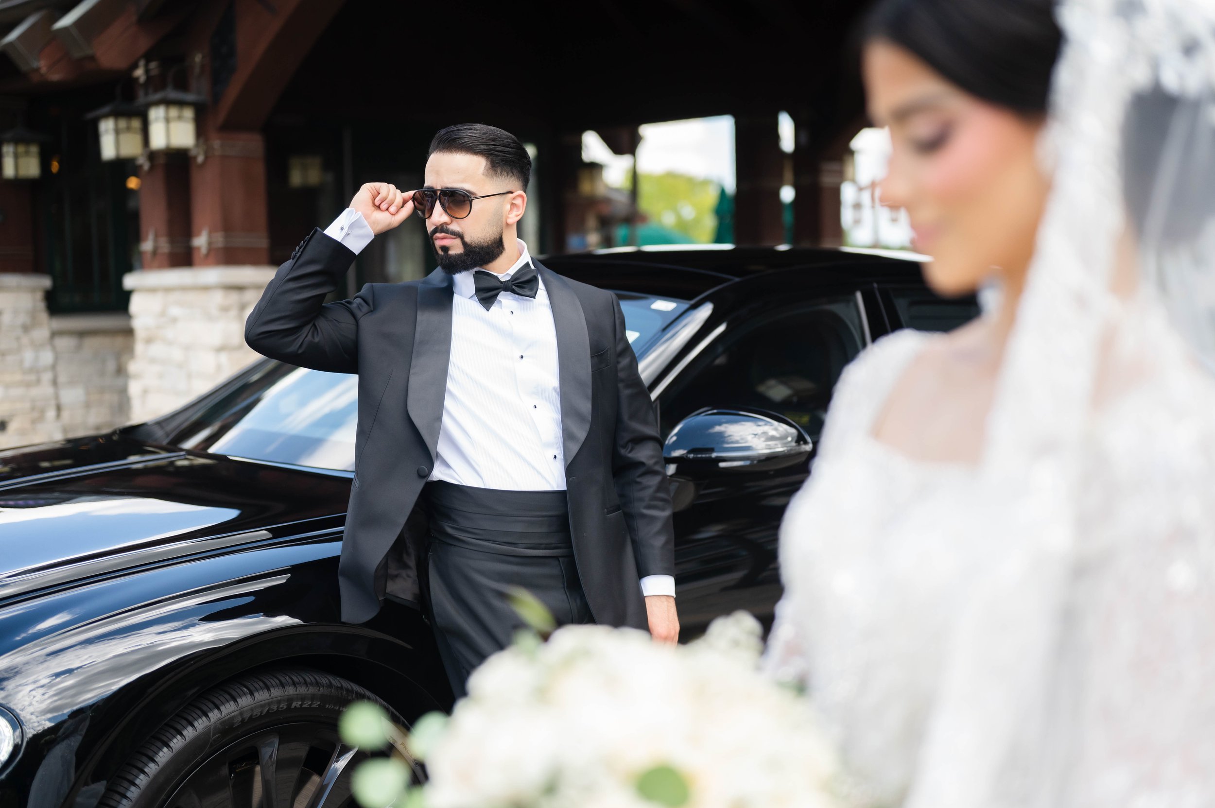 A man in a tuxedo and sunglasses stands near a black luxury car, looking to the side, while a woman in a wedding dress holds a bouquet in the foreground, slightly out of focus.