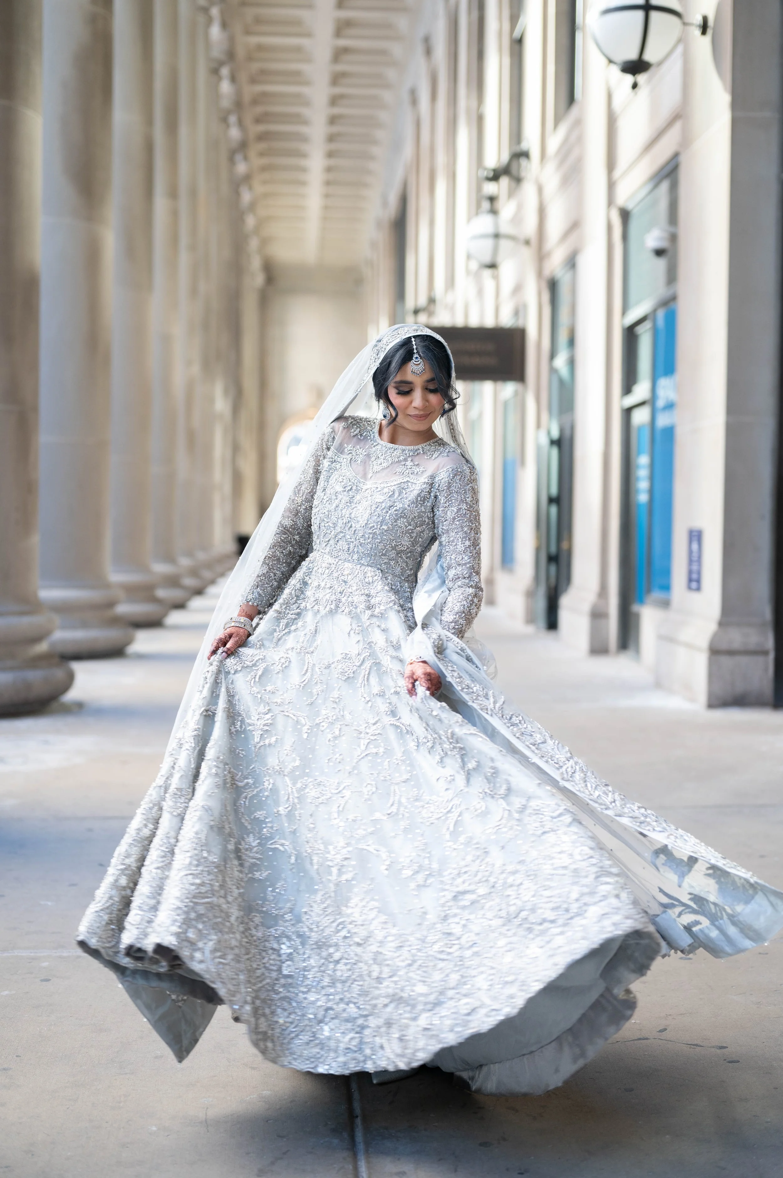 A bride in an elegant silver and white wedding gown with intricate embroidery, standing outdoors under a covered walkway with columns and hanging lights.