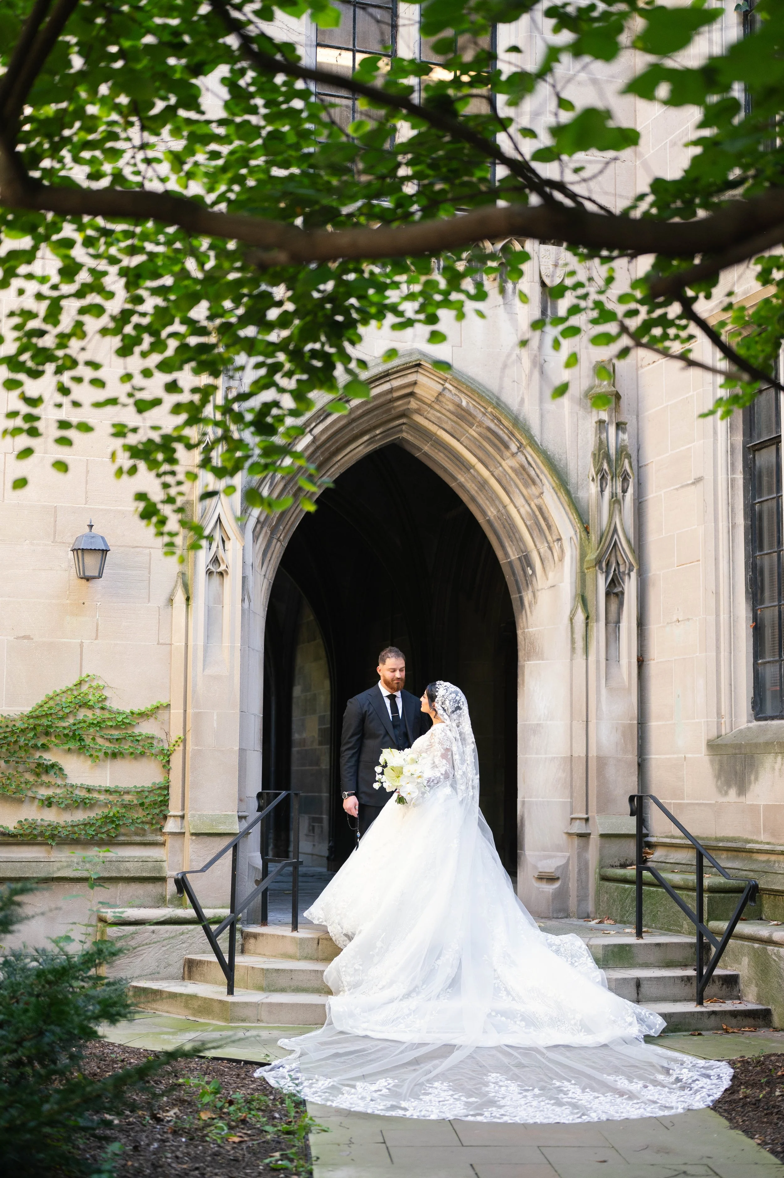 A bride and groom standing on the steps of a church, with the bride holding a bouquet of white flowers, and both dressed in wedding attire, framed by tree branches and leaves.