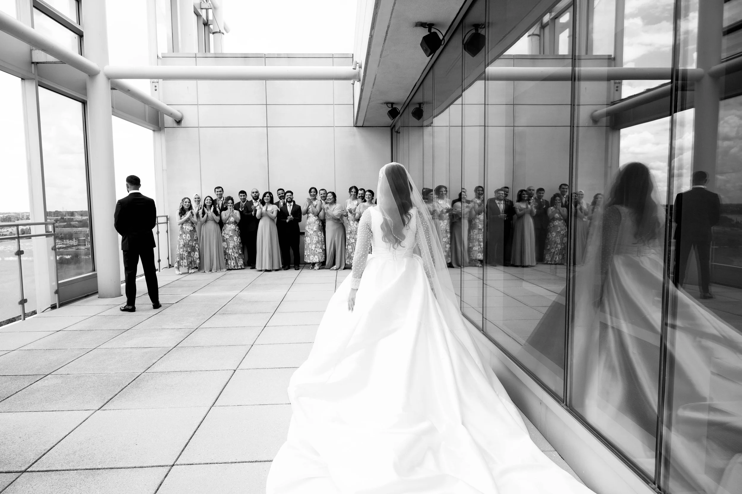 Bride in a wedding gown and veil looking at a group of bridesmaids and groomsmen on a rooftop terrace, with their reflections visible in glass windows.