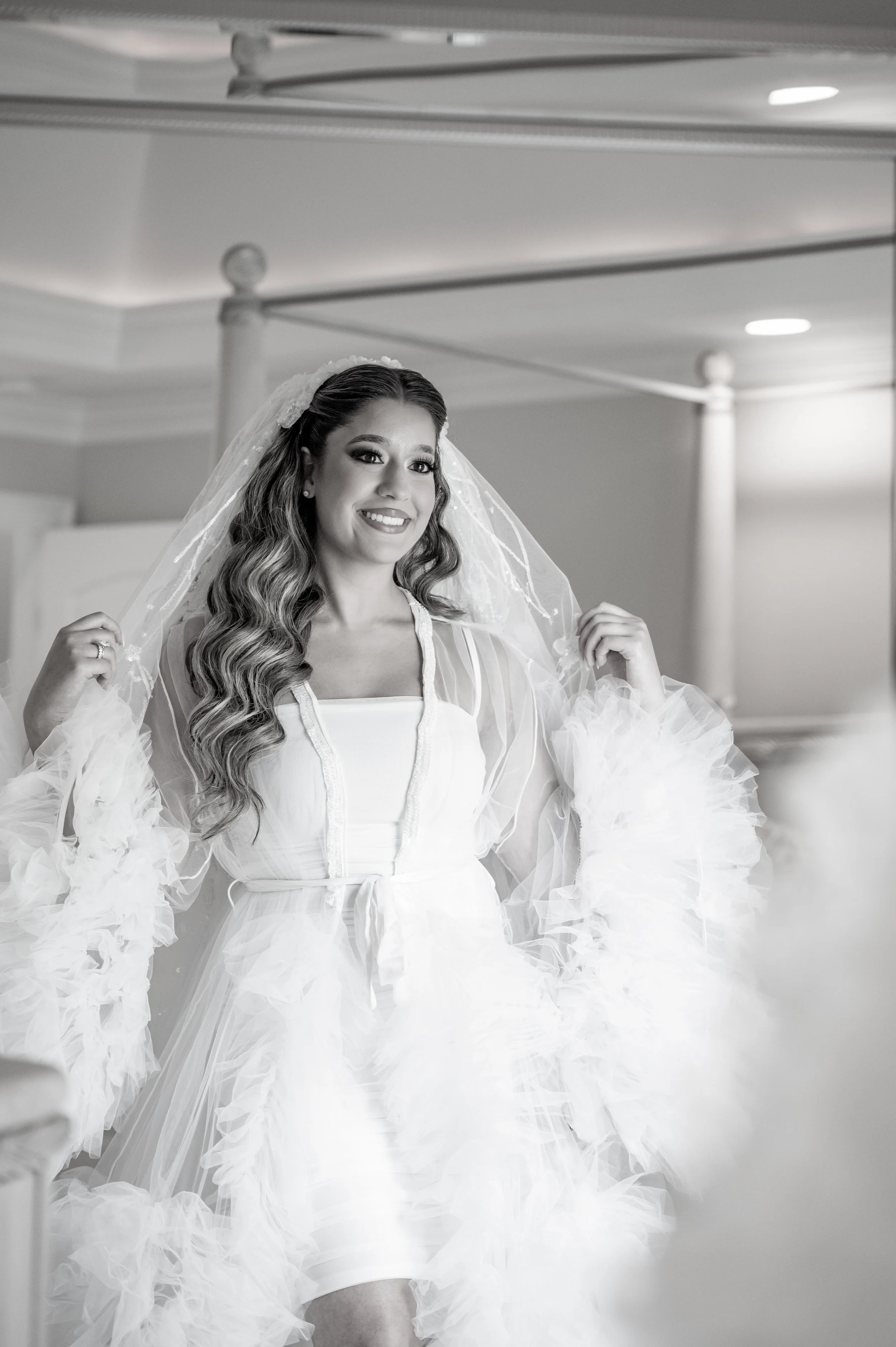 Black and white photo of a smiling bride in a wedding dress, holding her veil, in a room with a bed and columns.
