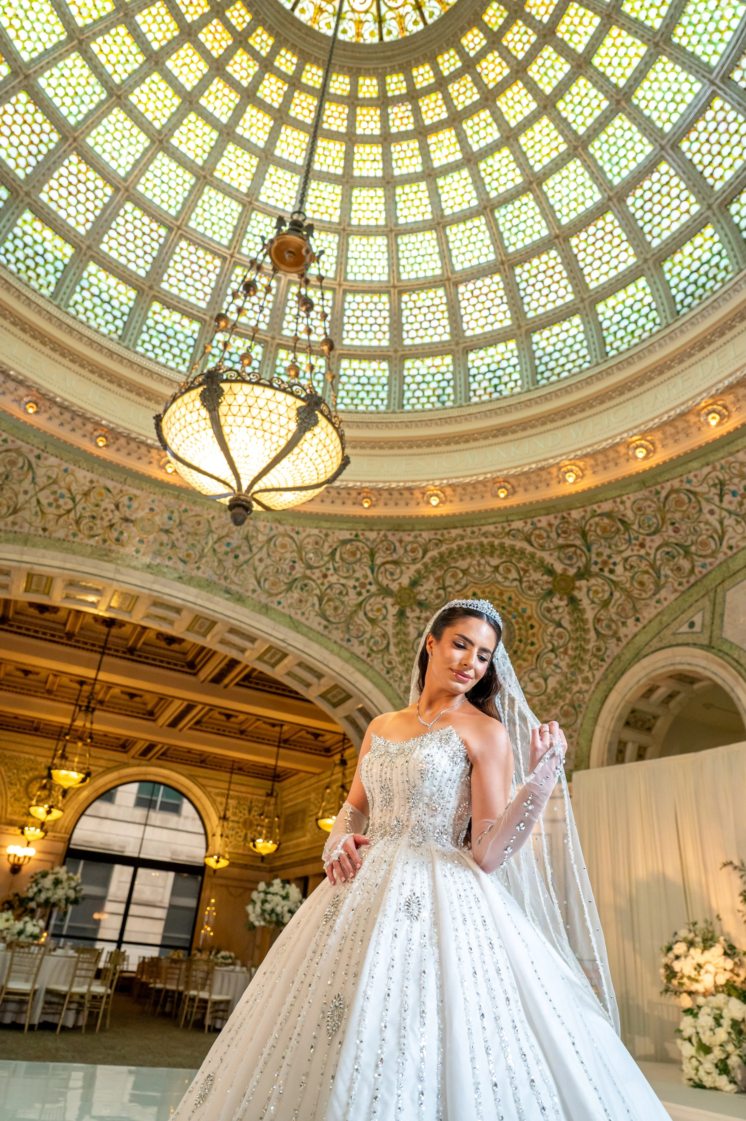 A bride in a wedding gown and veil standing inside a grand, ornate hall with a stained glass dome ceiling and chandeliers.