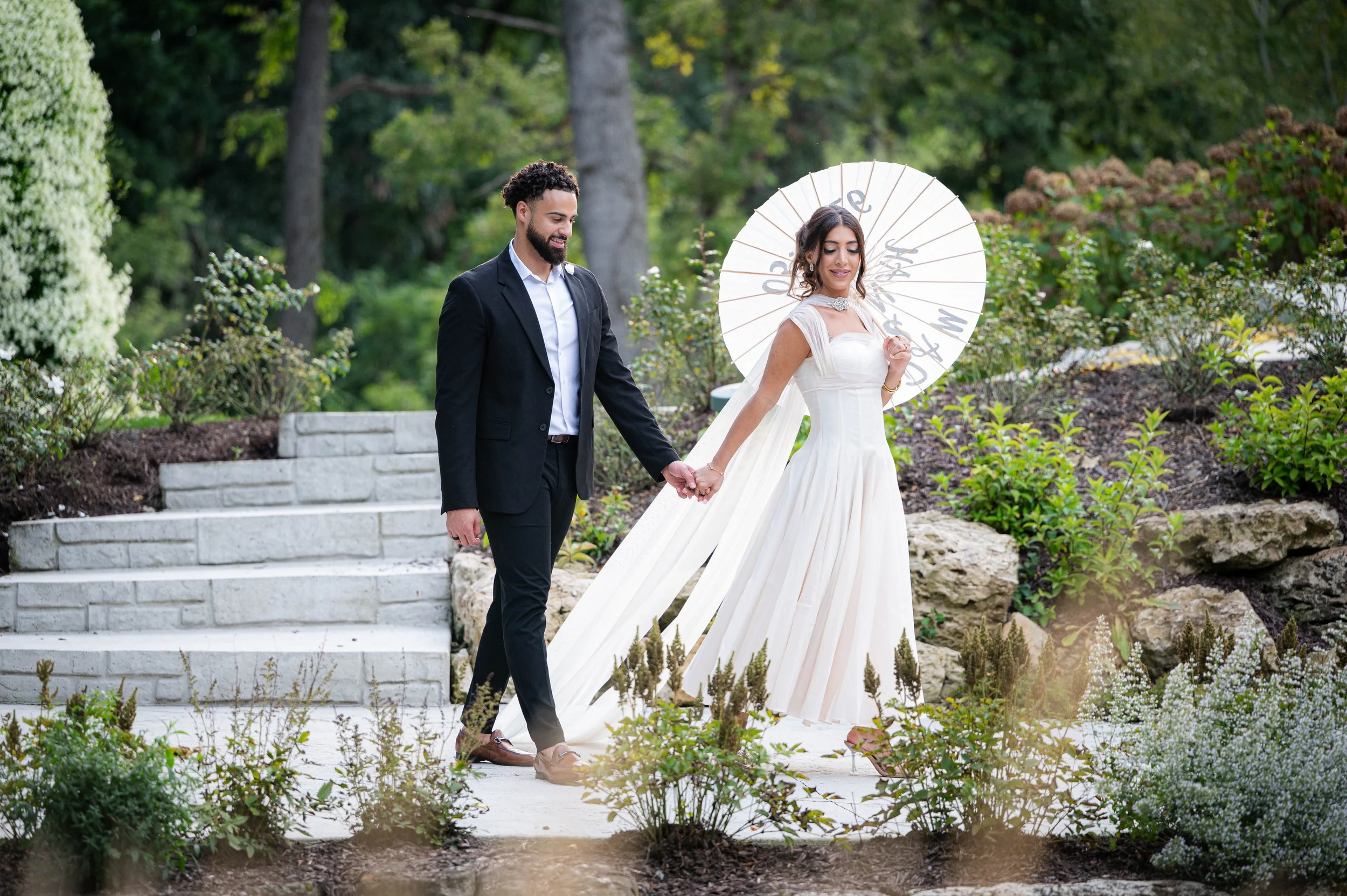 A bride and groom holding hands, walking on a stone pathway outdoors. The bride is dressed in a white gown and holding a paper parasol with writing. The groom is wearing a black suit and white shirt. They are surrounded by greenery and flowers.
