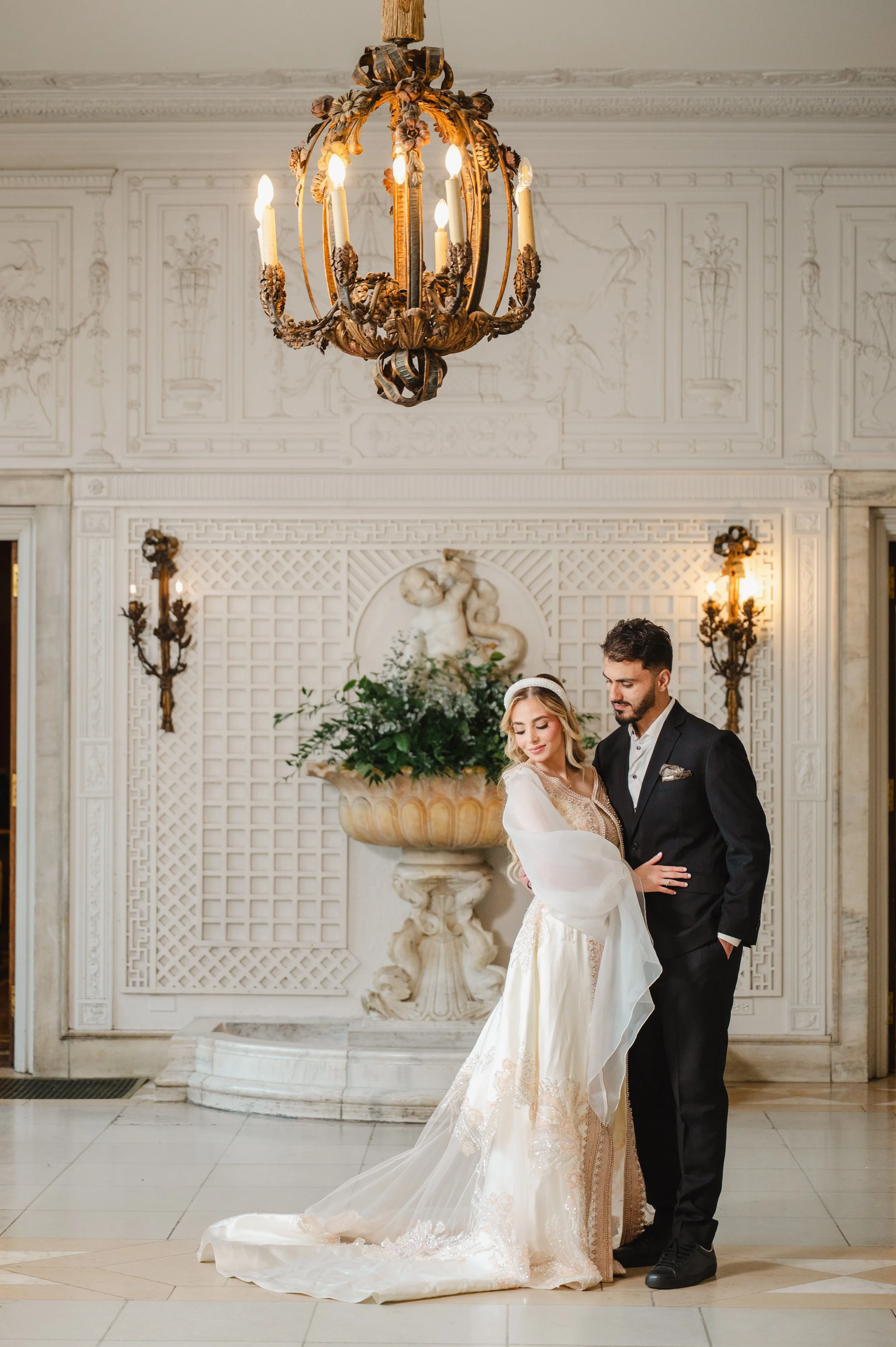 A bride and groom dancing in an elegant, ornate room with a chandelier, wall sconces, and a decorative fountain featuring a statue and greenery.