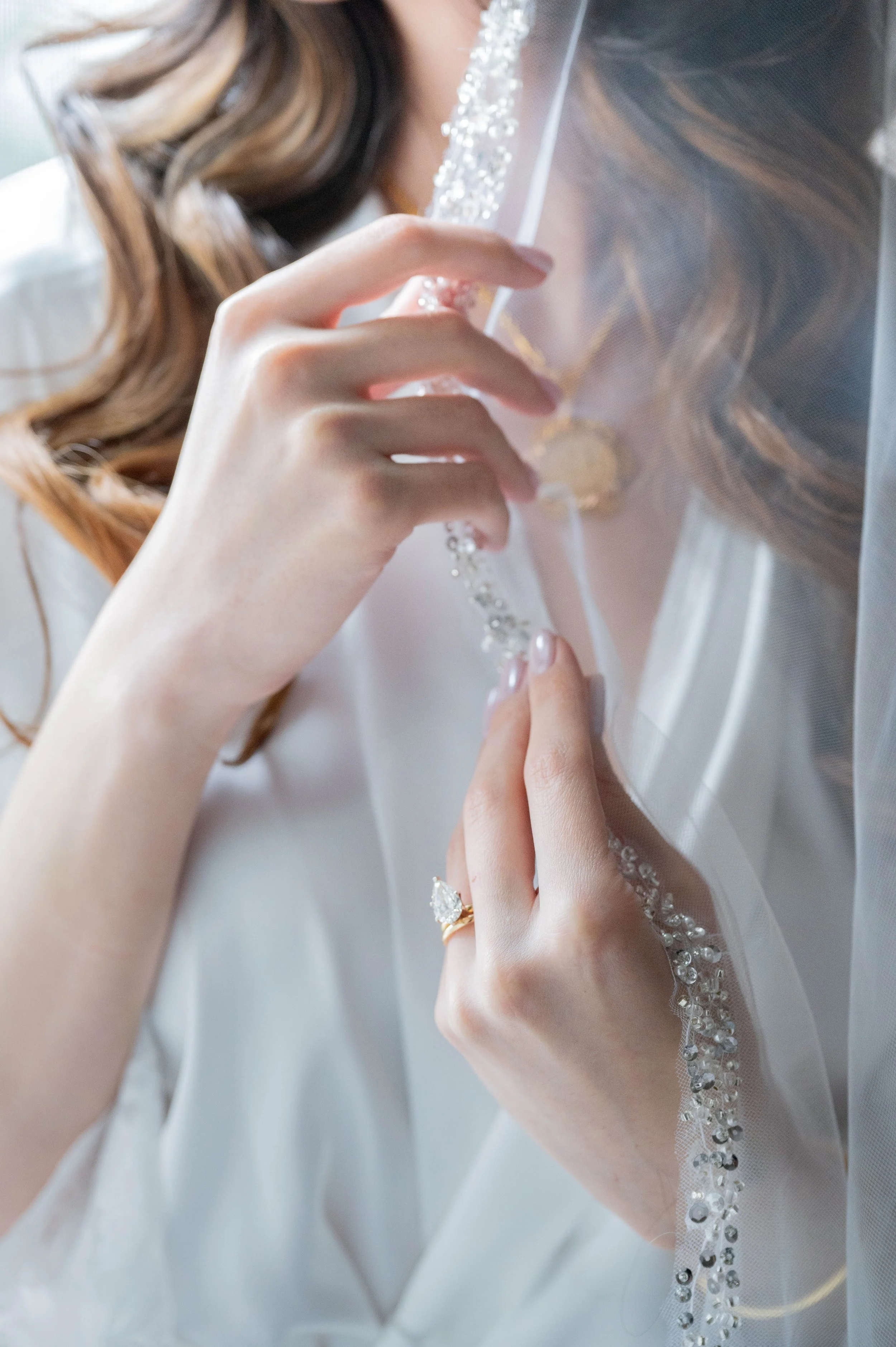 A woman in a white satin robe adjusts a sheer, beaded bridal veil, with her hand showing a gold ring with a large diamond.