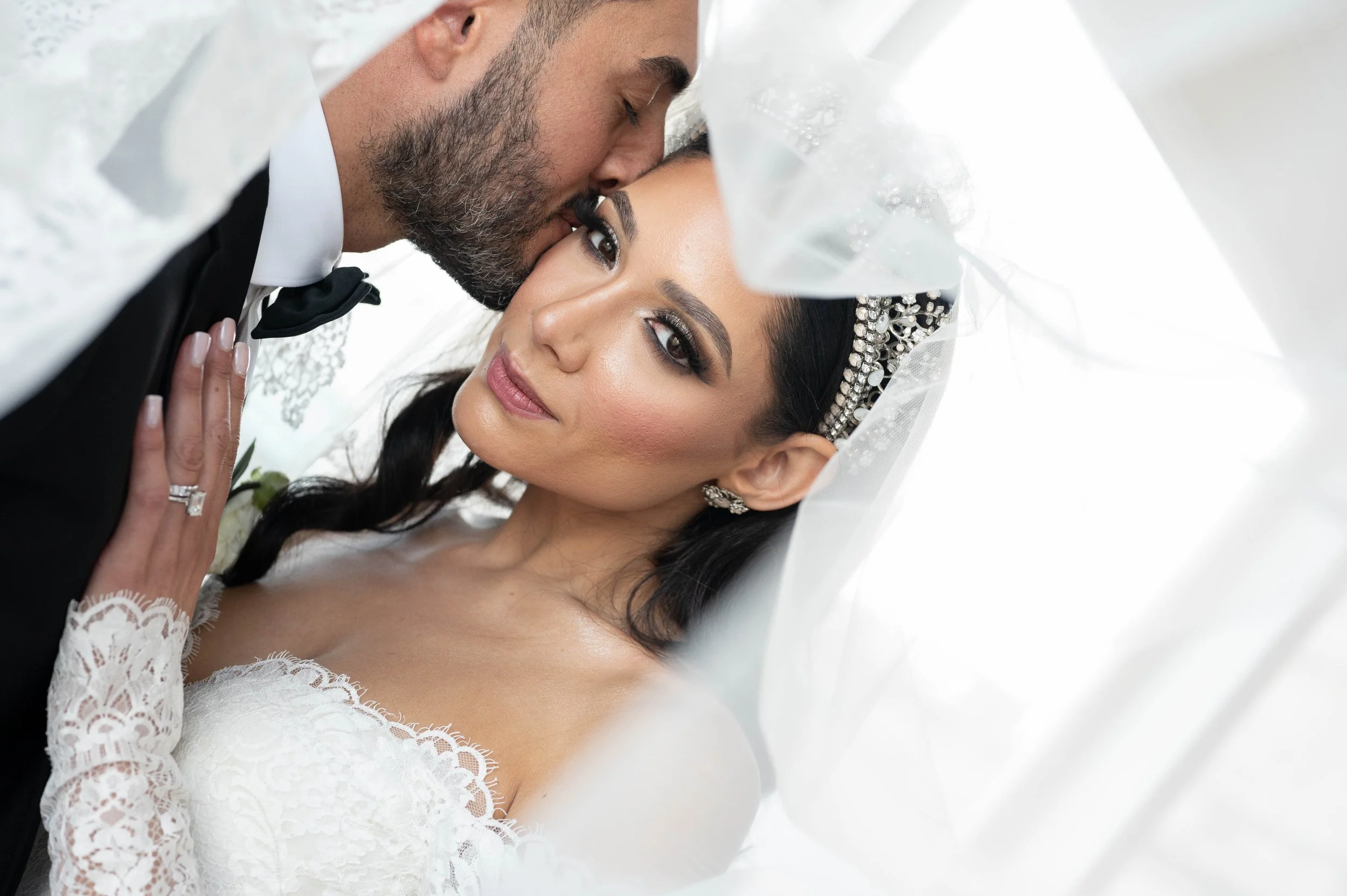 Close-up of a bride and groom in wedding attire, with the groom kissing the bride's forehead indoors.