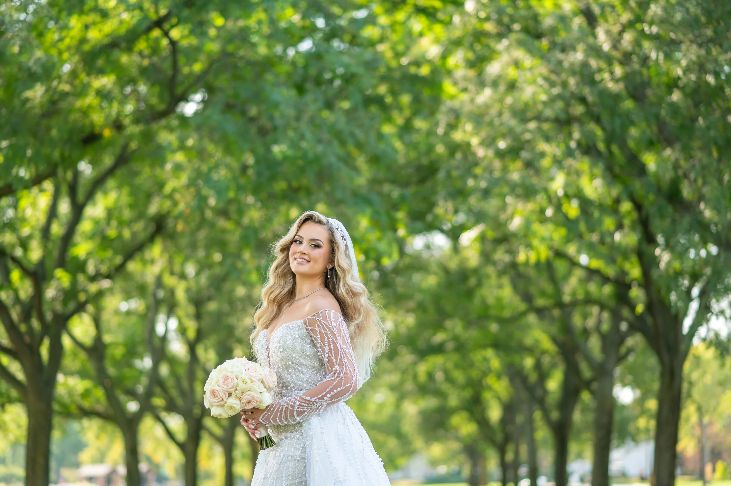 Bride in lace wedding dress holding bouquet of cream and pink roses standing outdoors under green trees.