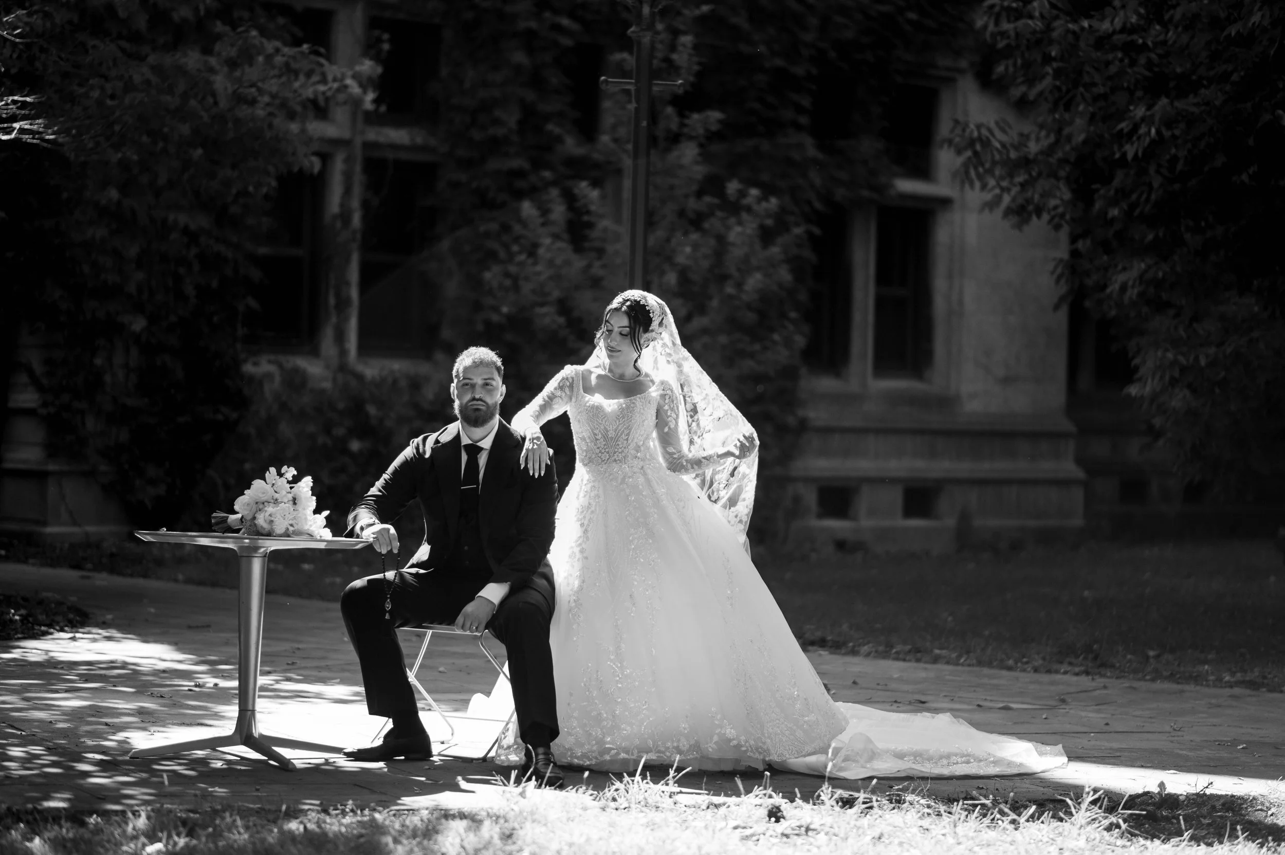 Black and white photo of a bride and groom outside, with the groom sitting and the bride standing beside him with her hand on his shoulder. The groom is wearing a suit and tie, and the bride is wearing a lace wedding dress with a long train and veil.