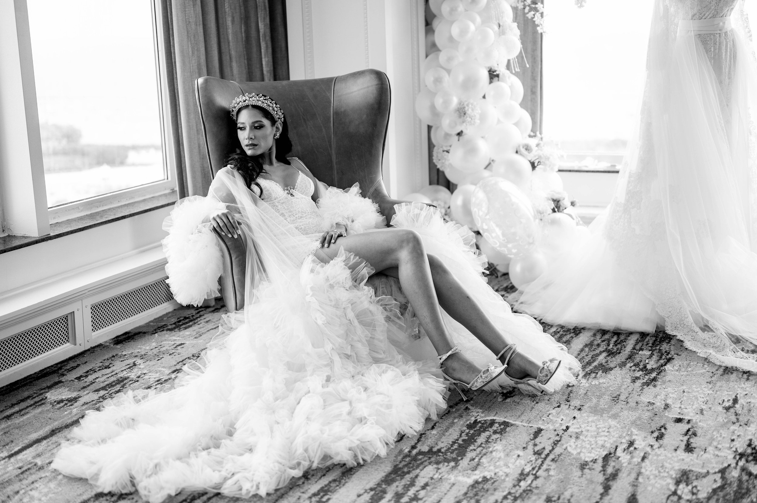 A woman in a wedding dress sitting on a high-back chair near a window, with balloons and a wedding gown in the background.