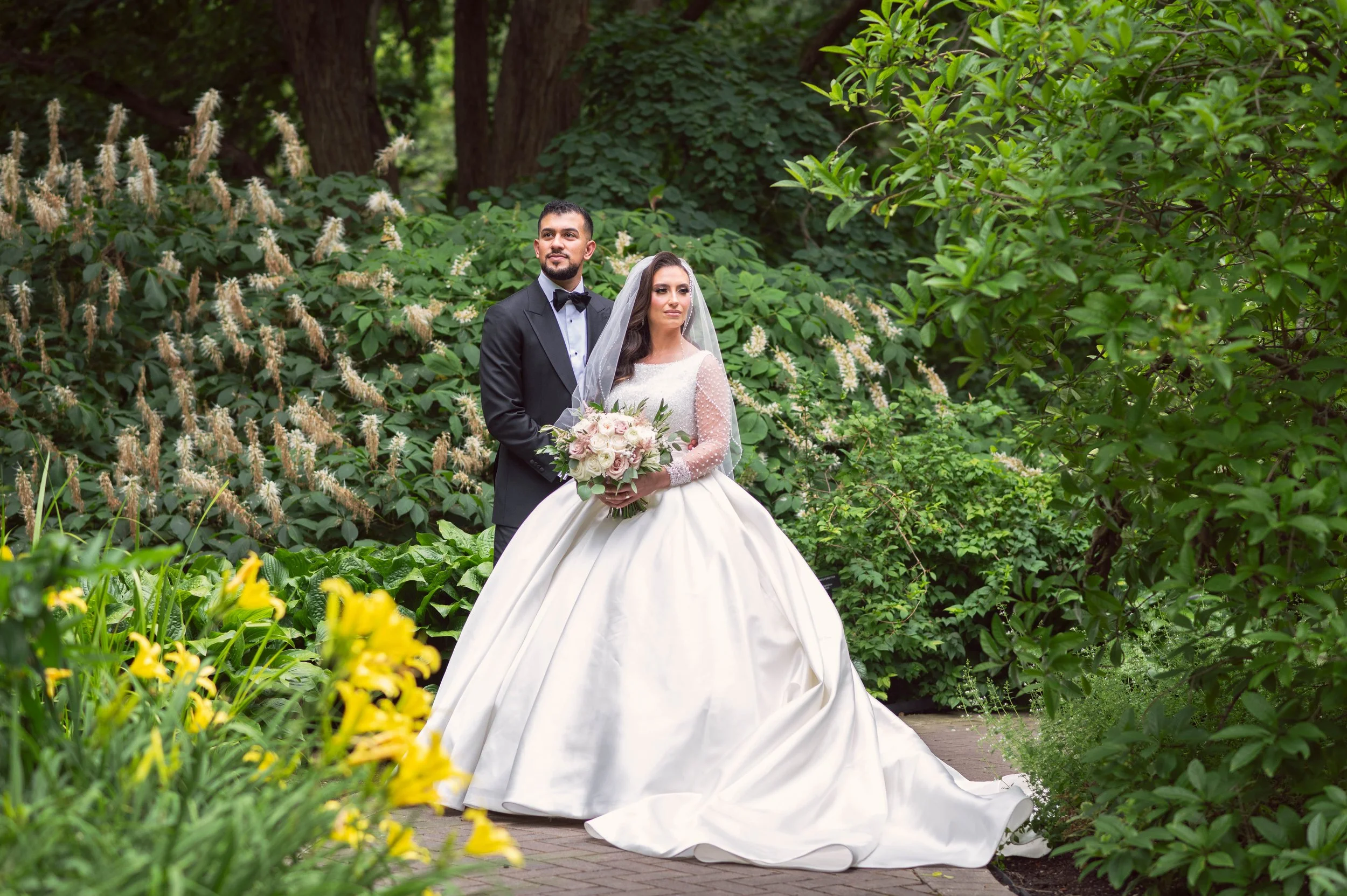 A bride and groom standing together in a lush, green garden. The bride is wearing a white wedding gown with a full skirt, lace sleeves, and a veil, holding a bouquet of pink and white flowers. The groom is dressed in a black tuxedo with a bow tie. Th