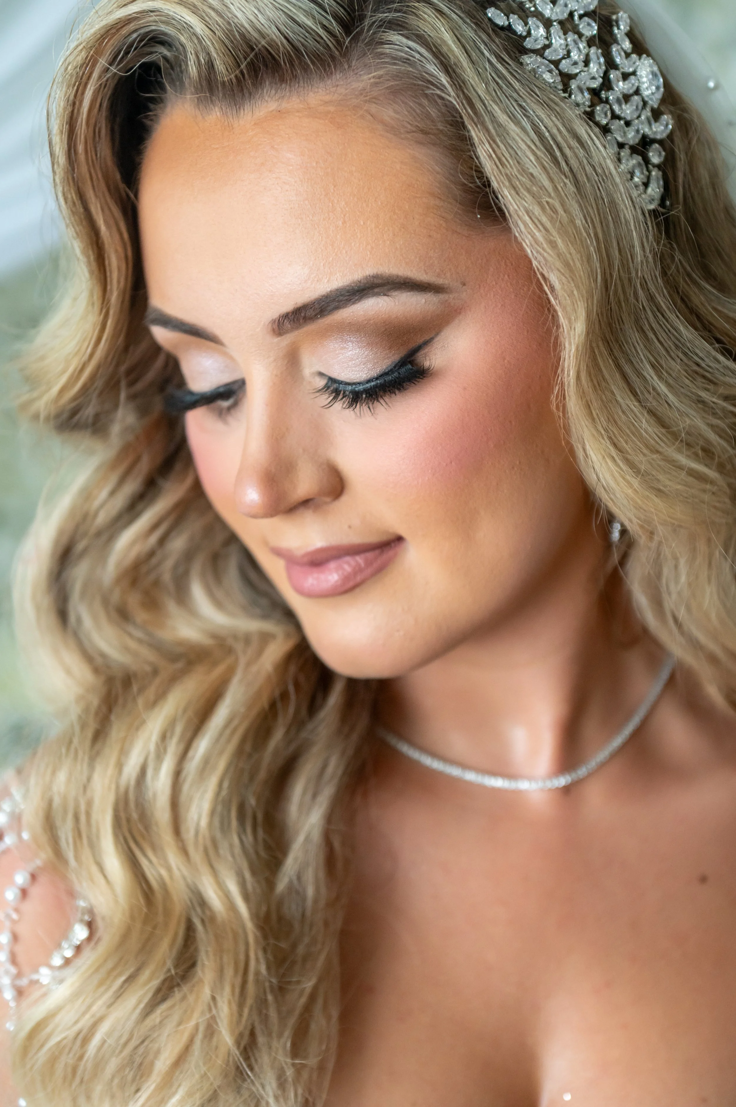 A woman with long blonde wavy hair, closed eyes, makeup, and a silver necklace, wearing a jeweled headband, looking downward.