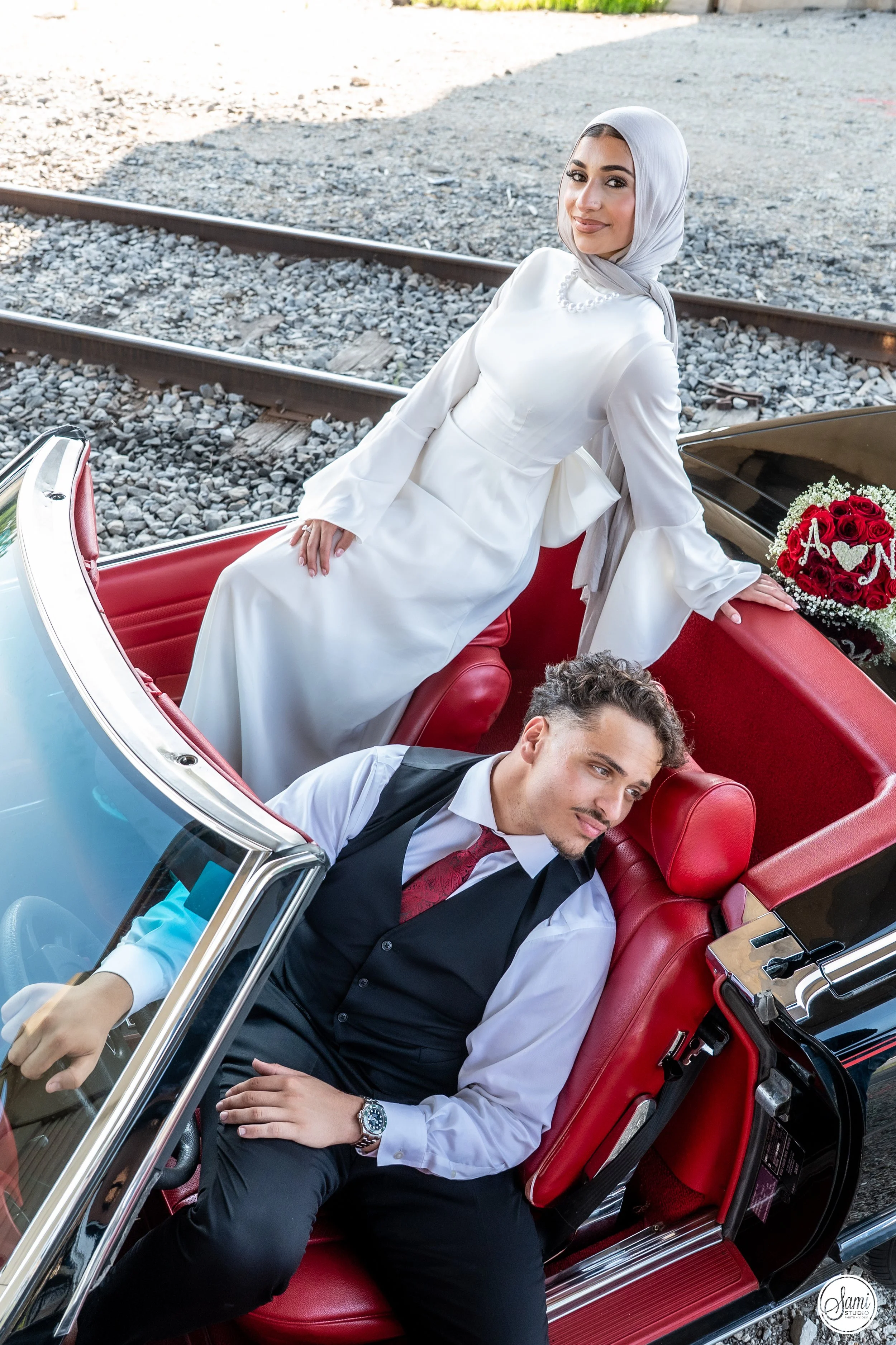 A woman in a white dress and grey hijab sitting in a vintage car, with a man in a black vest, white shirt, and red tie seated beside her. They are near train tracks, and there is a bouquet of red roses with a wedding sign on top of the car.