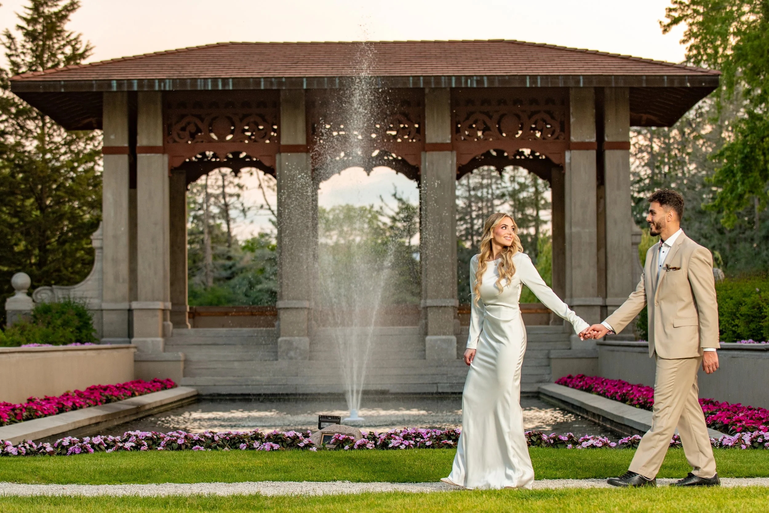 A bride and groom holding hands and walking in a garden with a fountain and a wooden gazebo in the background.