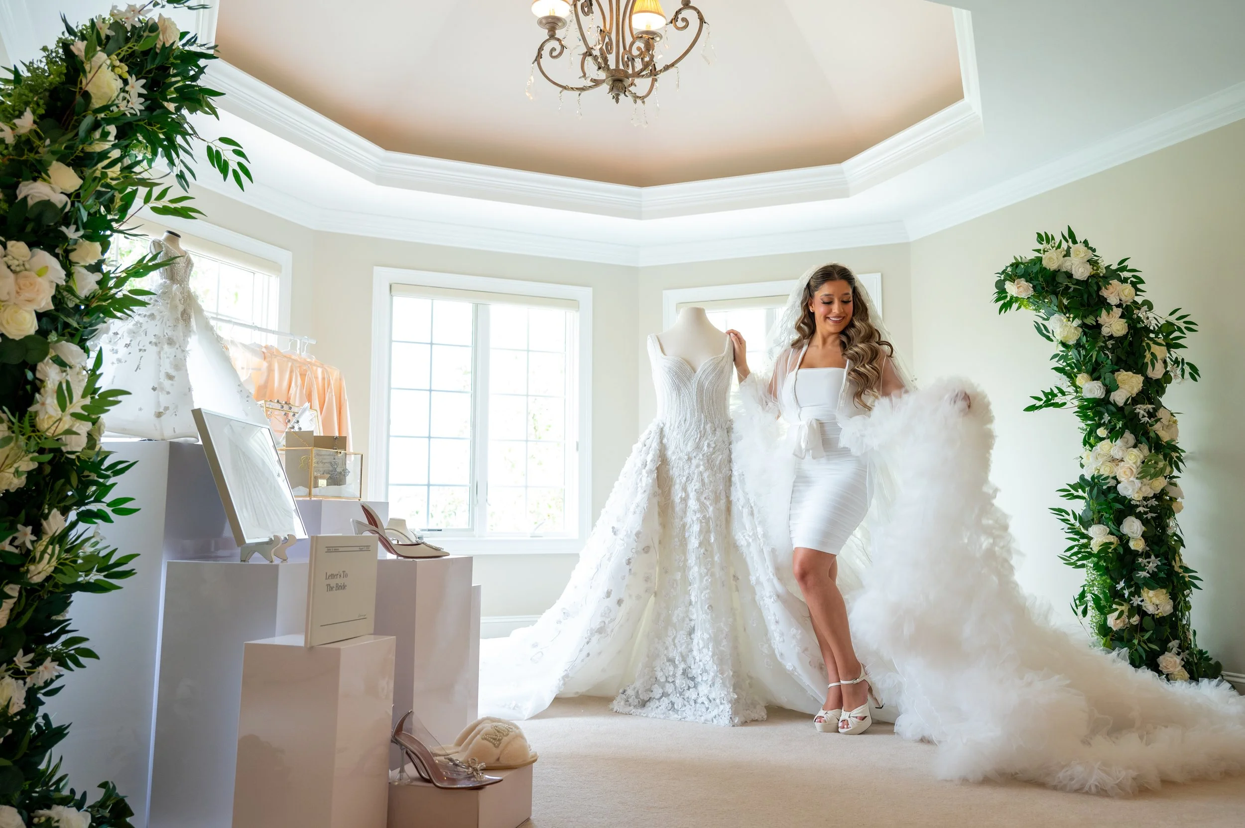 A bride with long, curly hair wearing a white dress and high heels, standing in a room decorated with floral arch, looking at a wedding gown on a mannequin, with a smile.