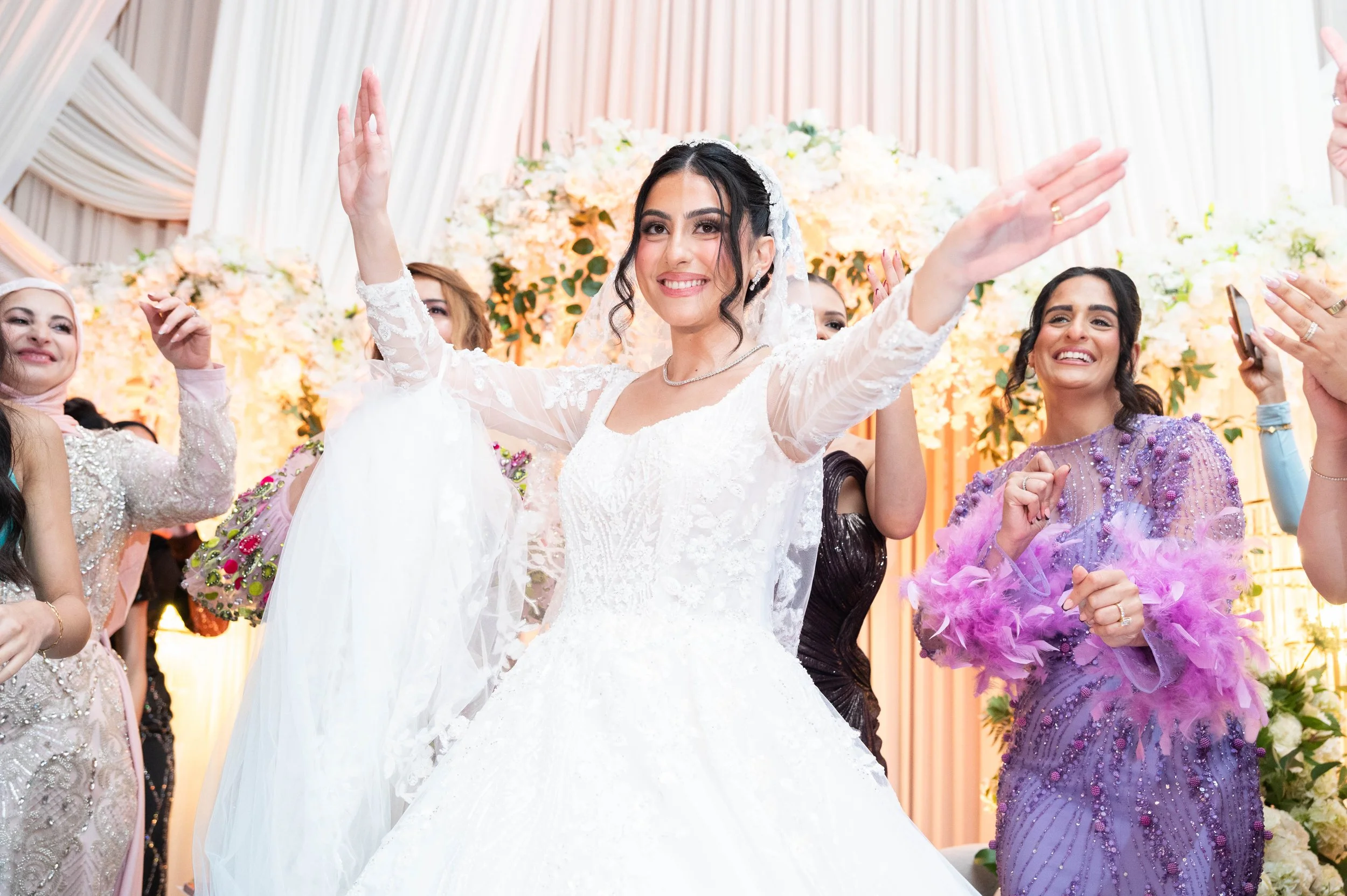 A bride in a white wedding gown smiling and celebrating with family and friends at her wedding reception, with floral decorations in the background.