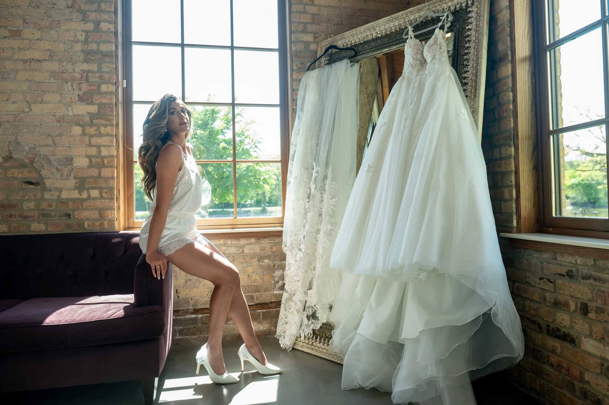 A woman in white satin lingerie with lace trim and white high heels sitting on a dark purple velvet sofa in a room with brick walls, large windows, and wedding dresses hanging on a mirror.