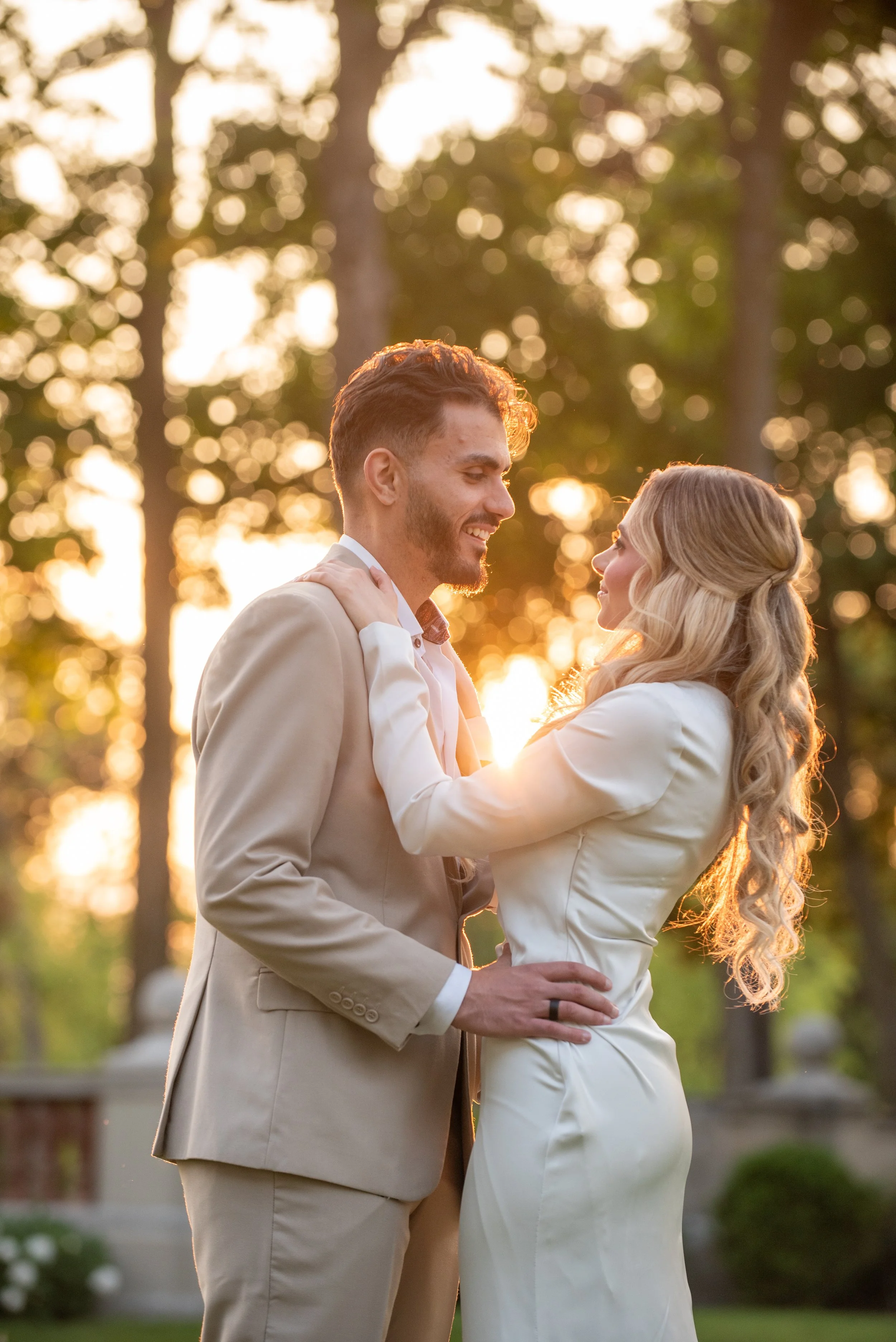 A couple dressed in wedding attire sharing a romantic moment outdoors at sunset, with trees and a stone railing in the background.